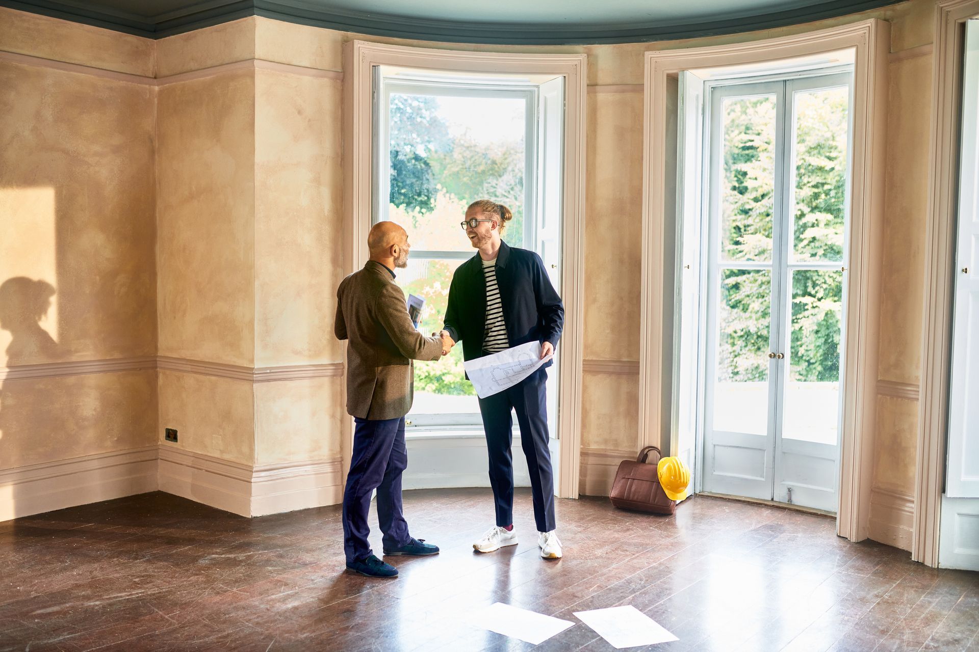 Two men shaking hands in a large, empty room with large windows, possibly a real estate deal.
