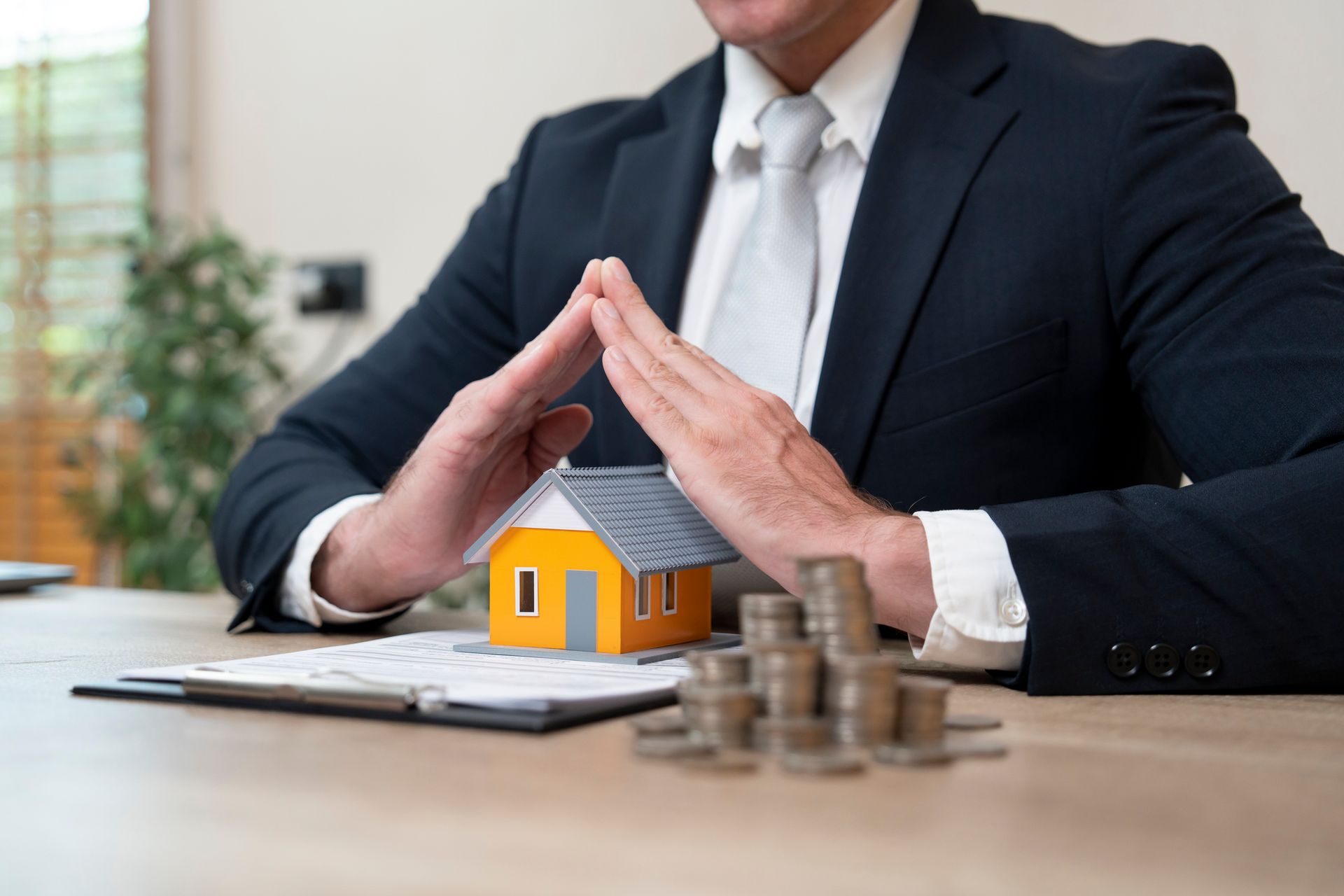 Person in a suit protecting a model house with hands, stacks of coins nearby, and papers on a desk.