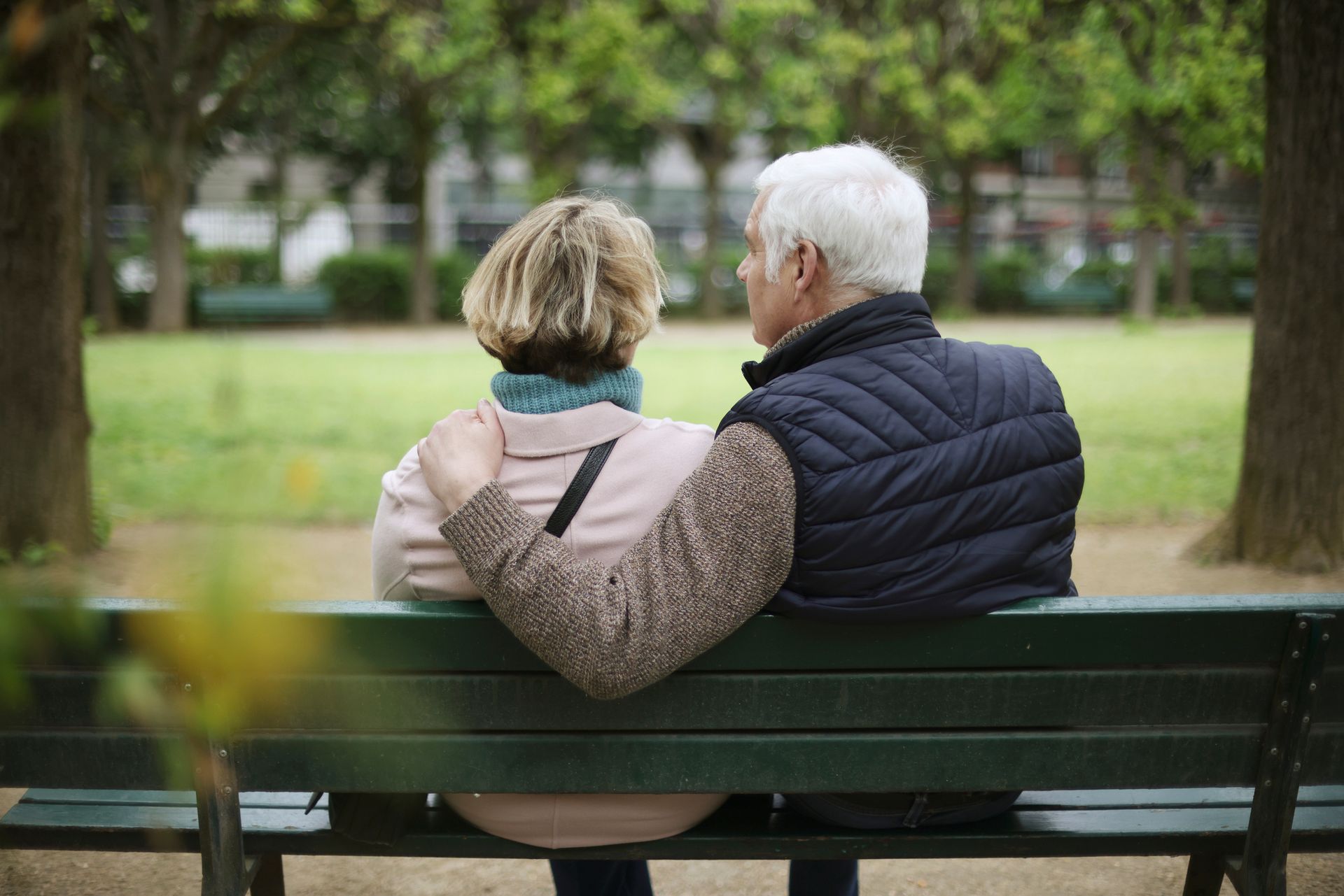 Older couple sitting on a park bench, arm around each other.