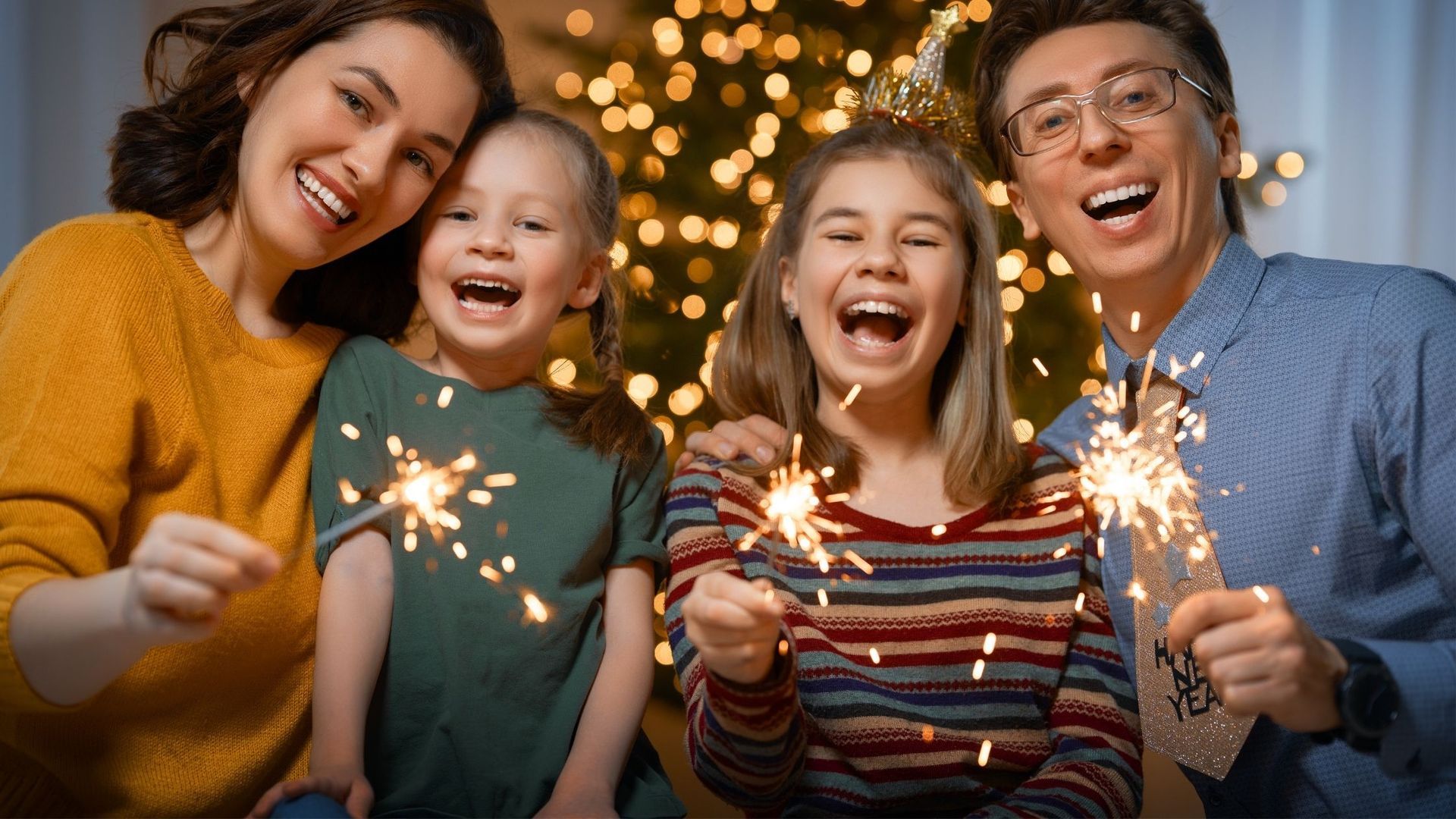 Family with sparklers smiling in front of a Christmas tree with golden lights.