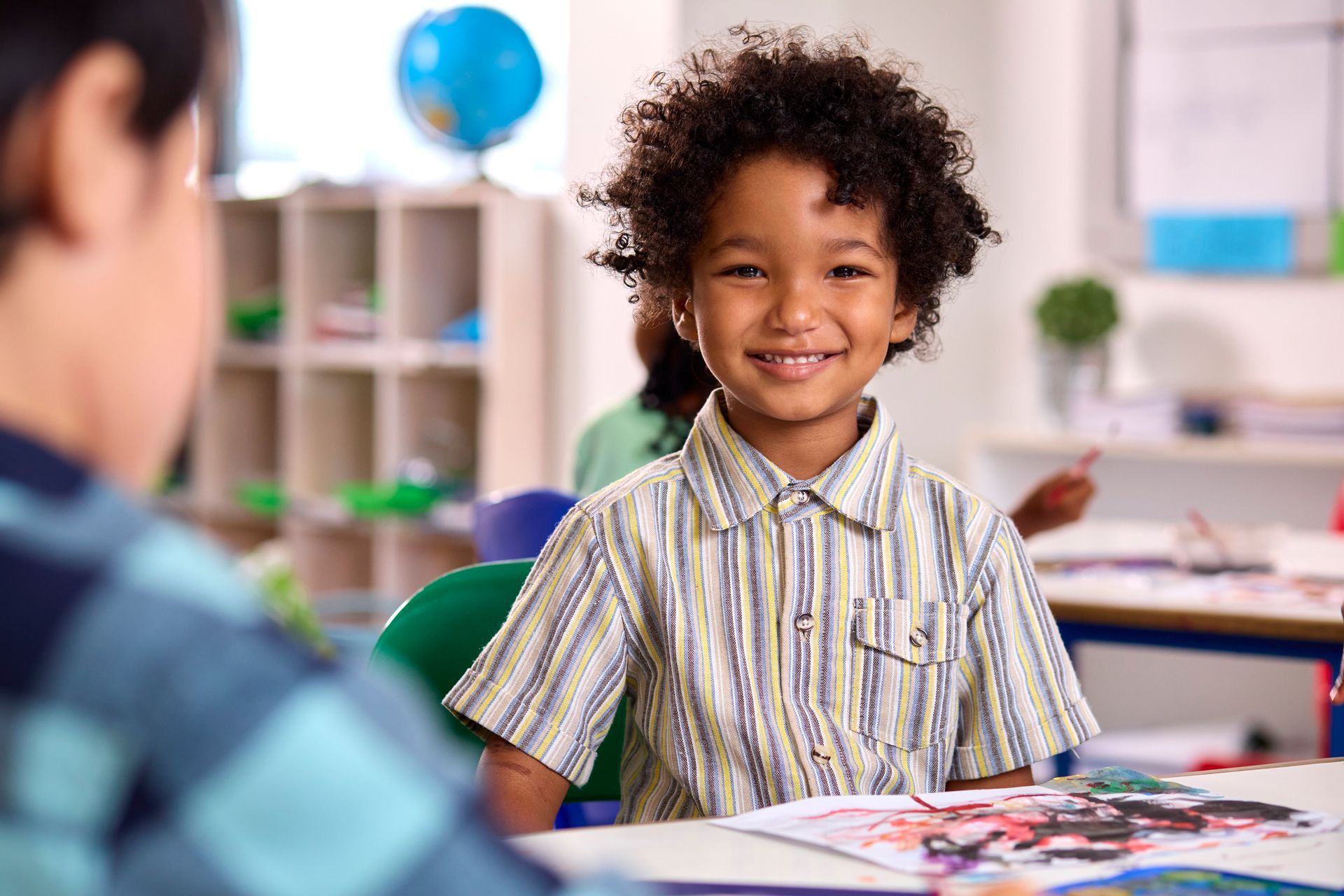 A young boy is smiling while sitting at a table in a classroom.