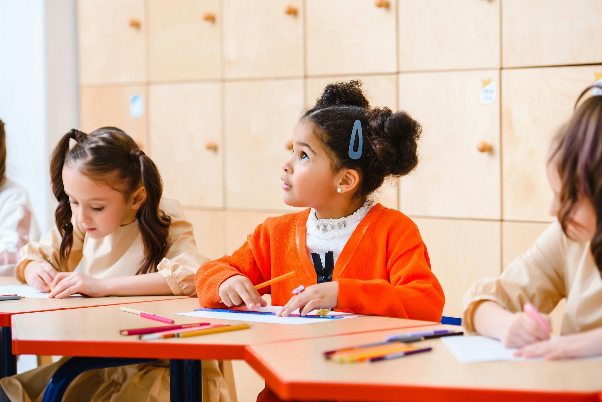 Children at desks in a classroom, drawing with colored pencils. Girl in orange cardigan looks up.
