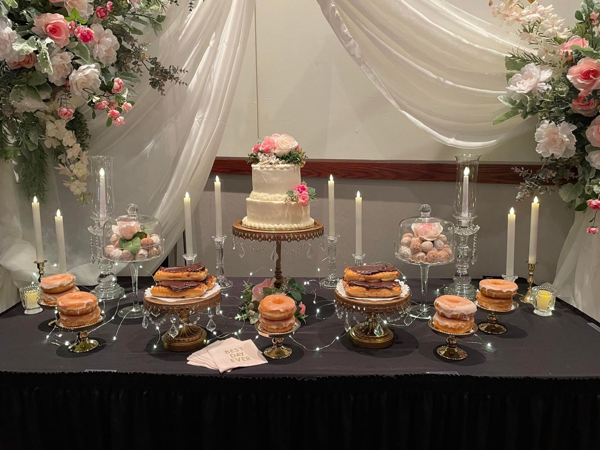 Elegant dessert table with tiered cake, pastries, candles, and pink floral arrangements under draped white fabric
