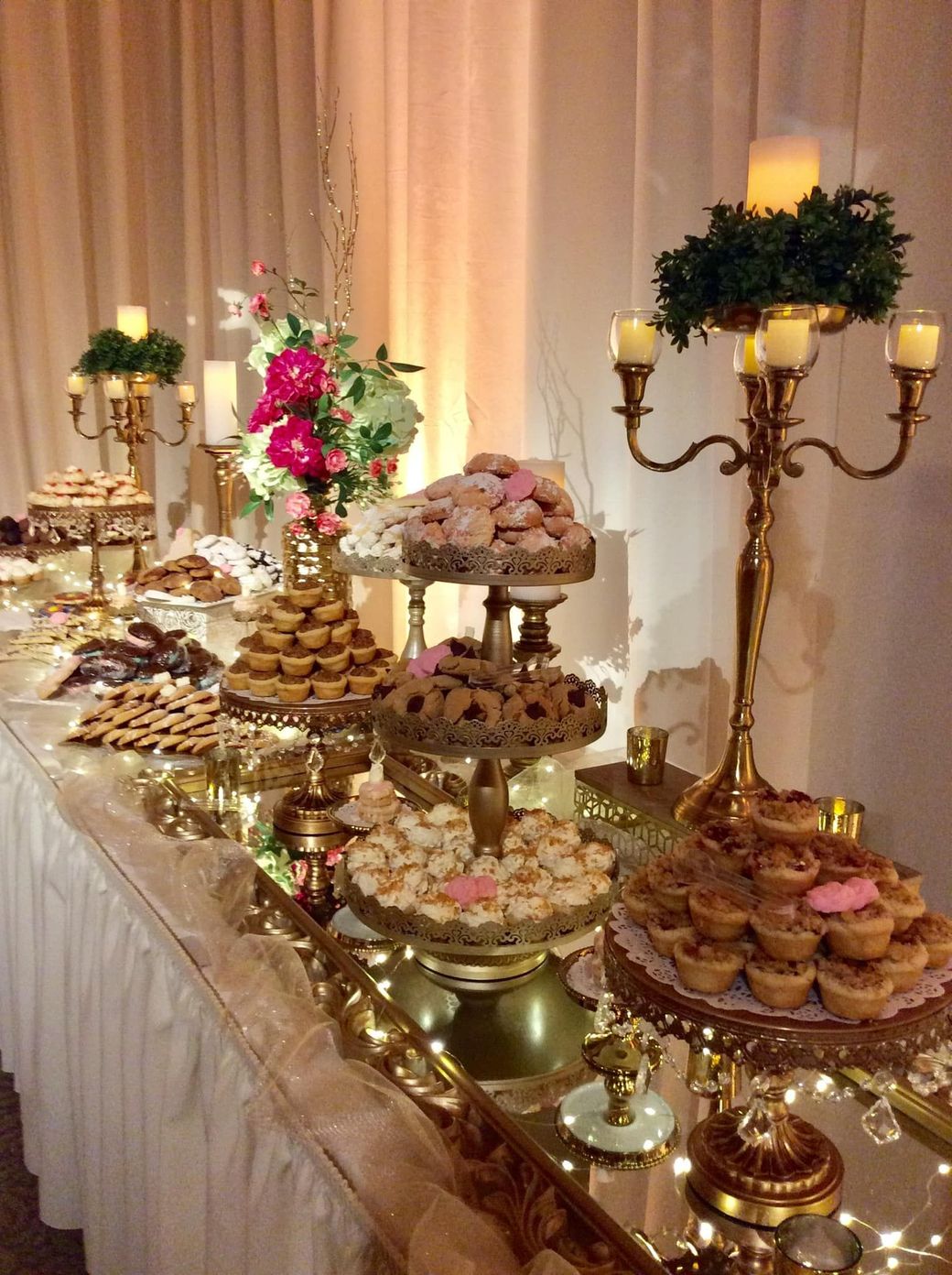 Elegant dessert buffet with gold candleholders, white drapery, and trays of pastries and flowers
