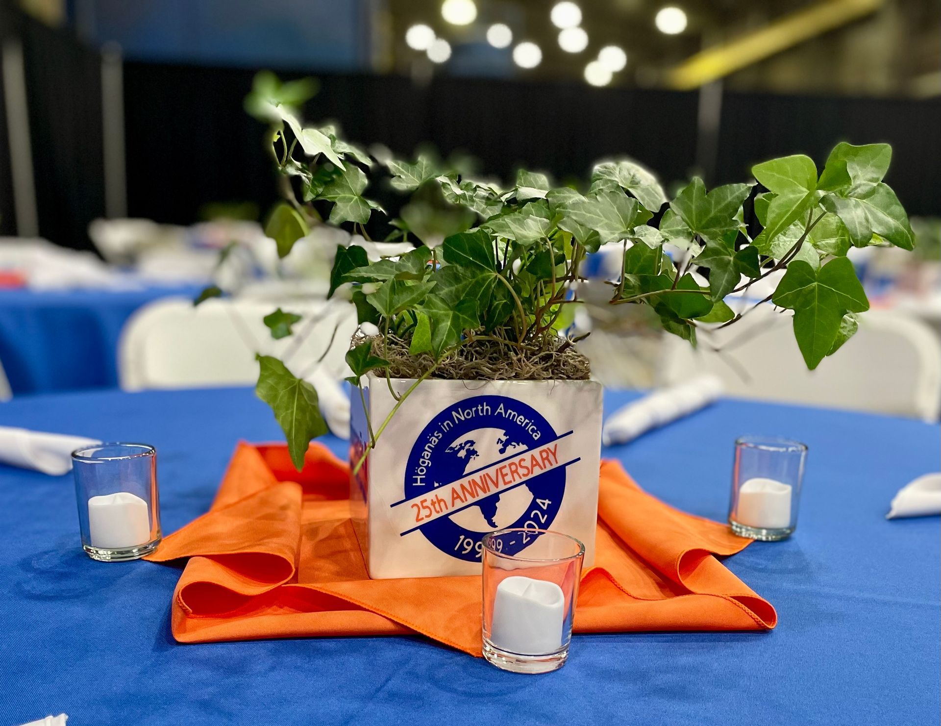 Table centerpiece with green plant in a glass pot on an orange napkin, surrounded by small candles on a blue tablecloth