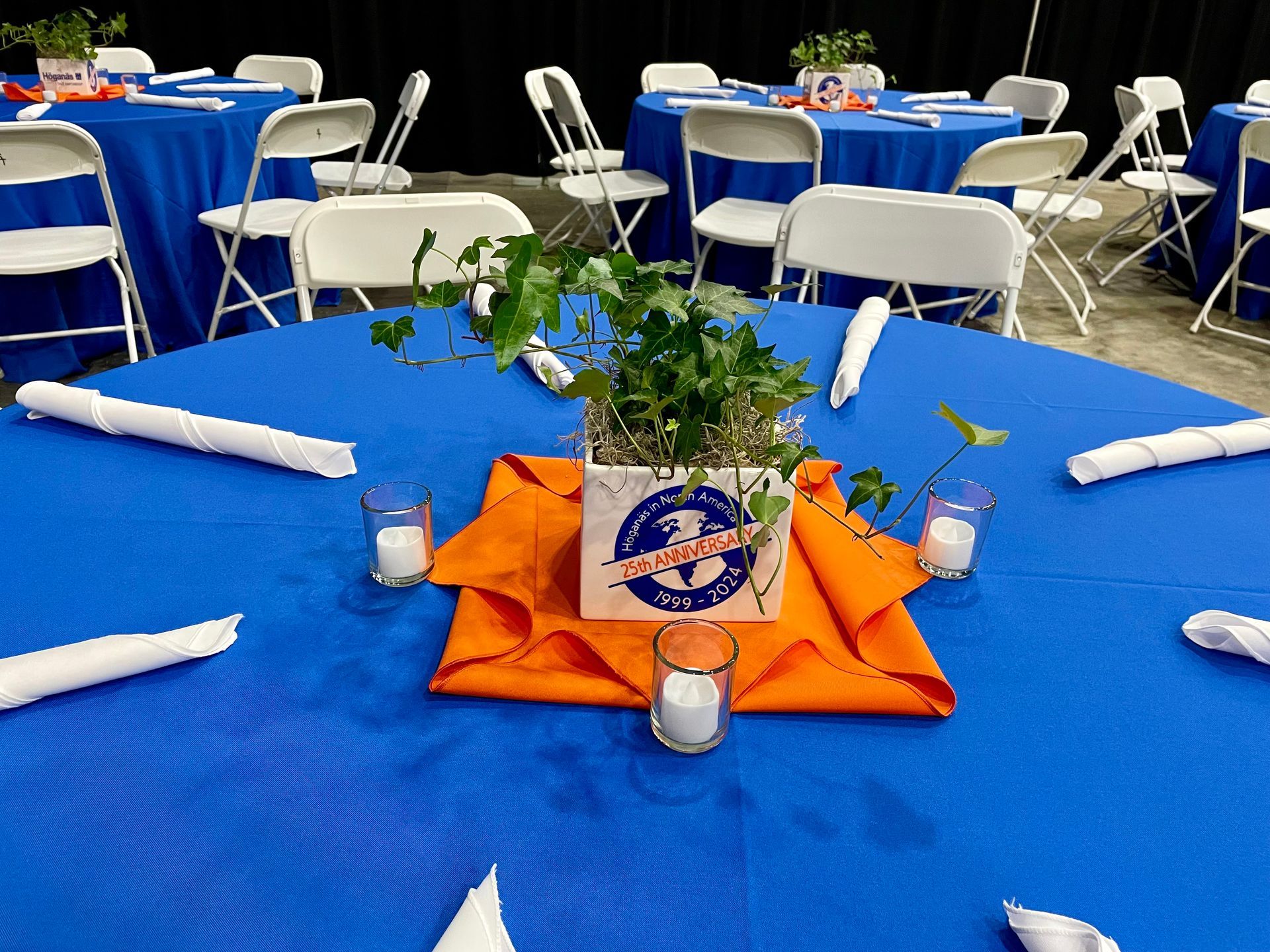 Blue banquet table with white chairs, folded napkins, candles, and a floral centerpiece