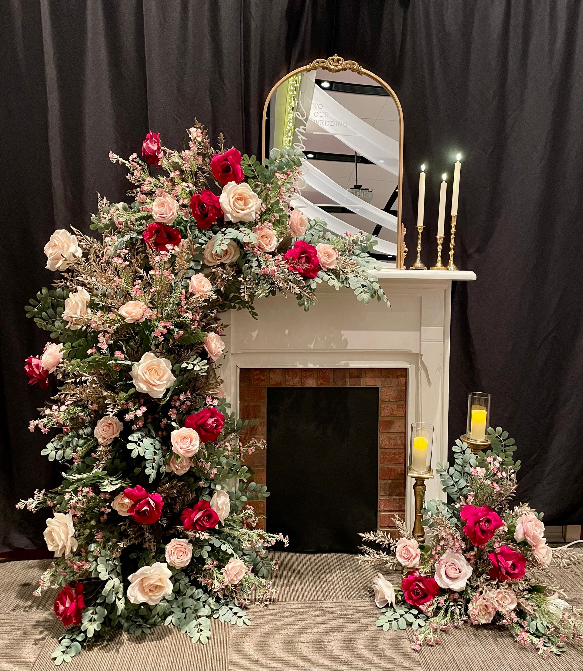 Floral-decorated fireplace display with pink and white roses, candles, mirror, and black drapery backdrop