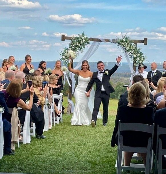 Newlyweds wave under a floral arch at an outdoor wedding ceremony with seated guests.