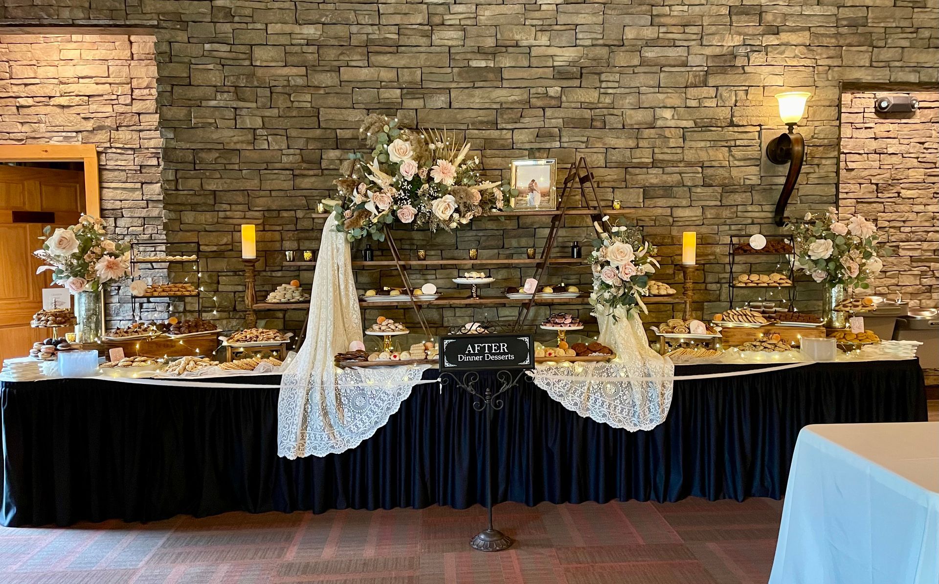 Stone-wall banquet table with white lace runners, flowers, candles, and gold accents at a rustic event