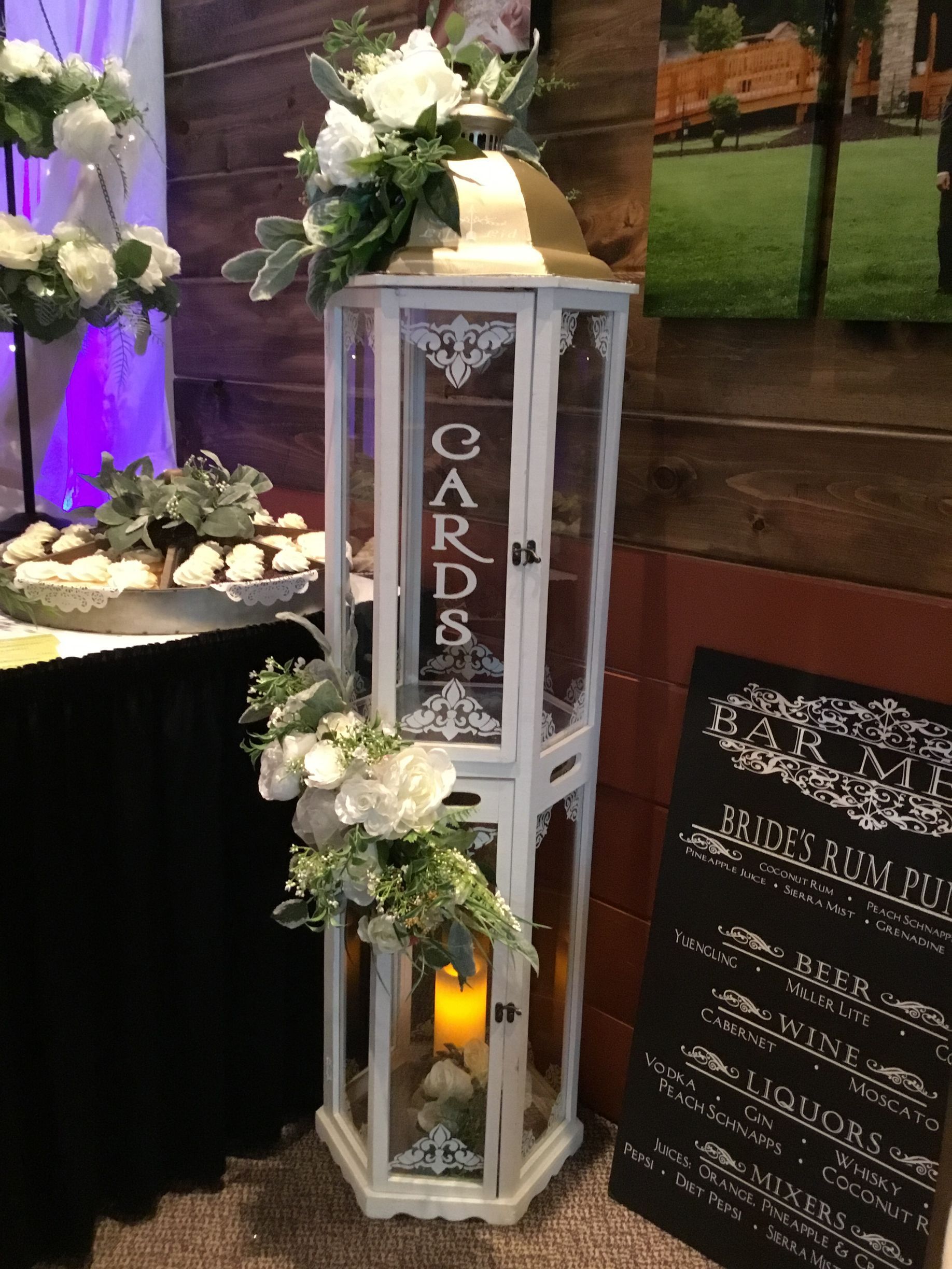 White lantern-style memorial stand with candle, white flowers, and a vertical “CARDS” sign indoors