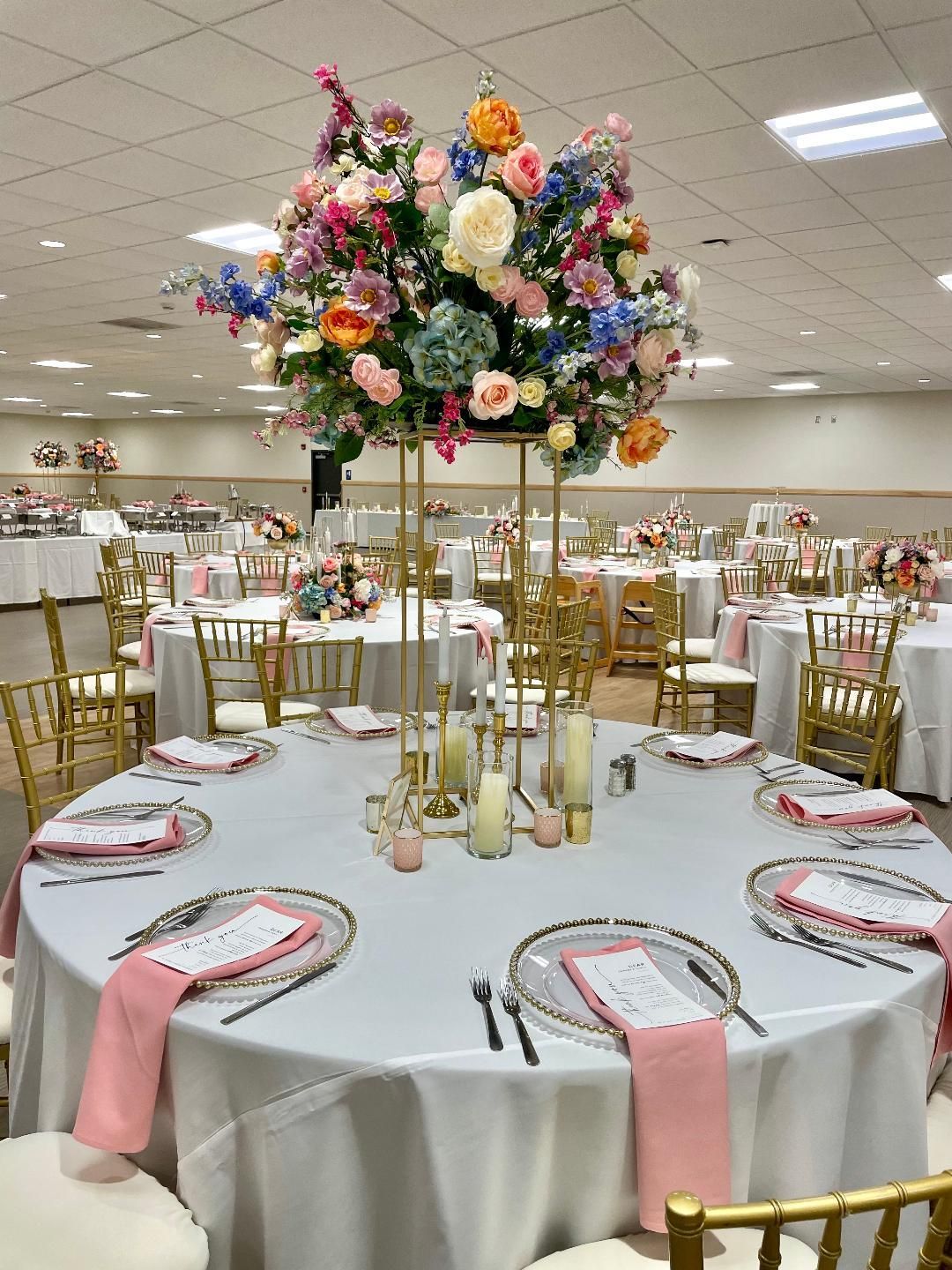 Round banquet table with white cloth, pink napkins, and tall floral centerpiece in a reception hall