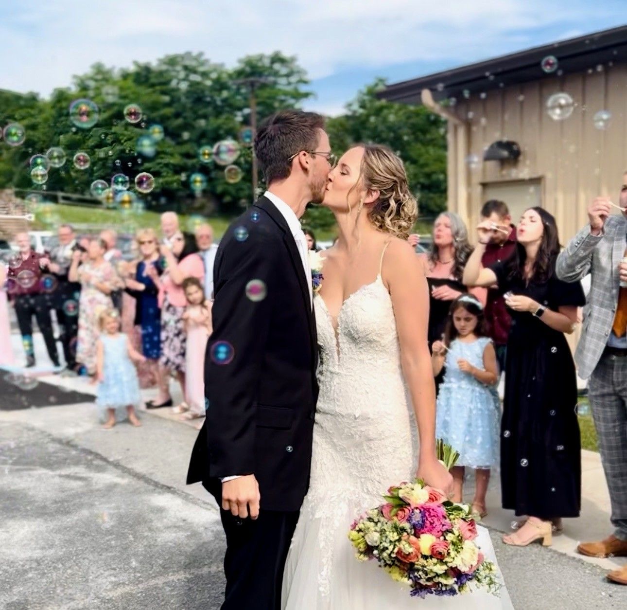 Bride and groom kiss at an outdoor wedding with guests and bubbles in the background