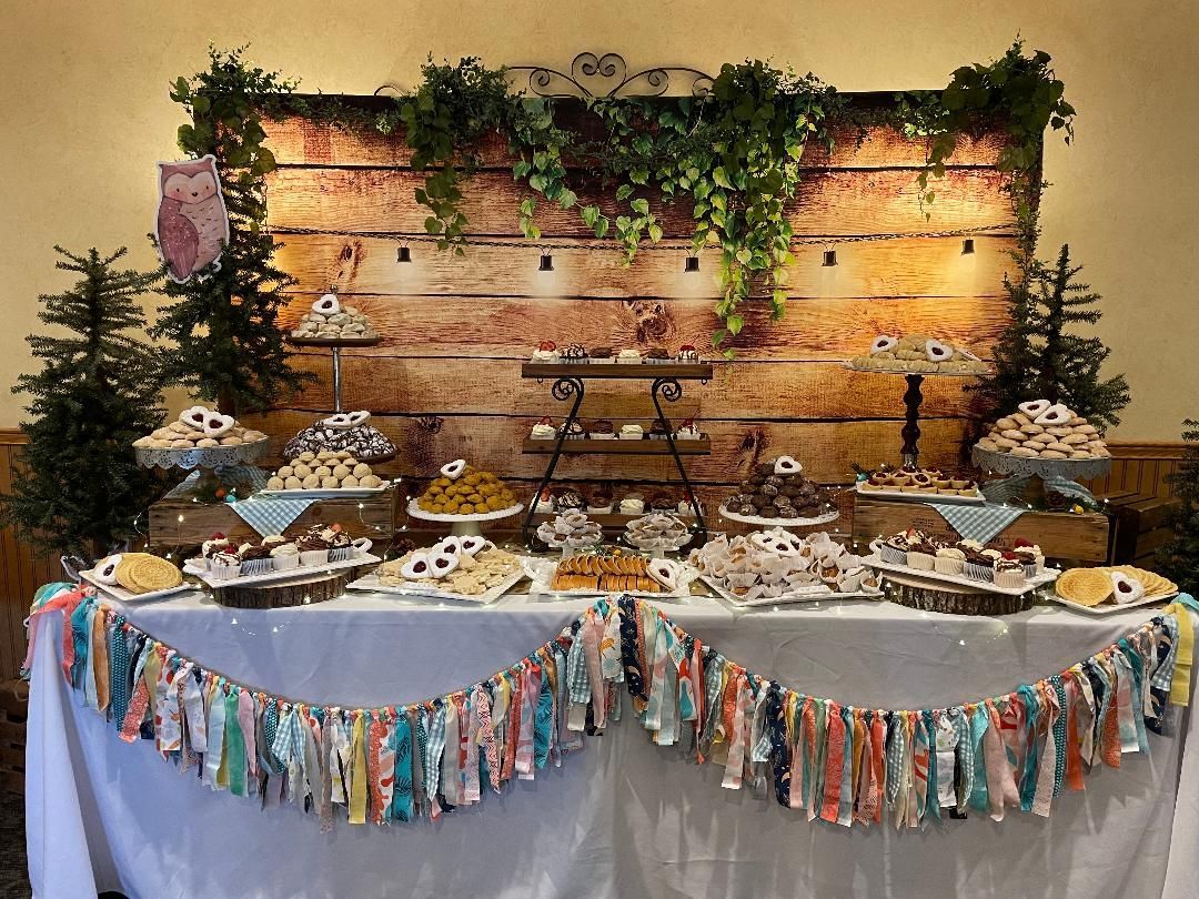 Buffet table with desserts and greenery against a rustic wooden backdrop, decorated with colorful fringe trim
