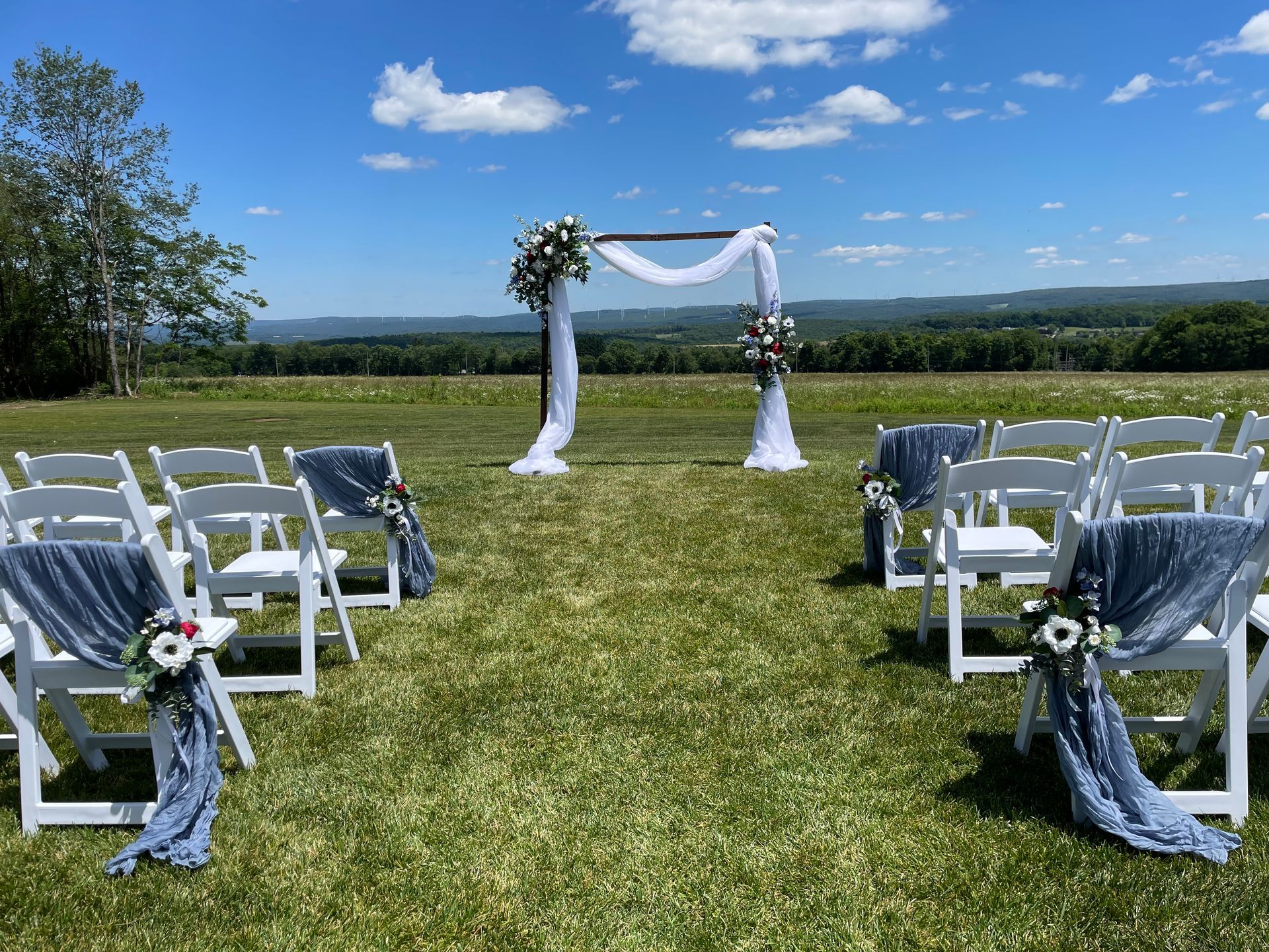 Outdoor wedding ceremony setup with white chairs and blue drapes facing a flower-decorated arch on a grassy field.