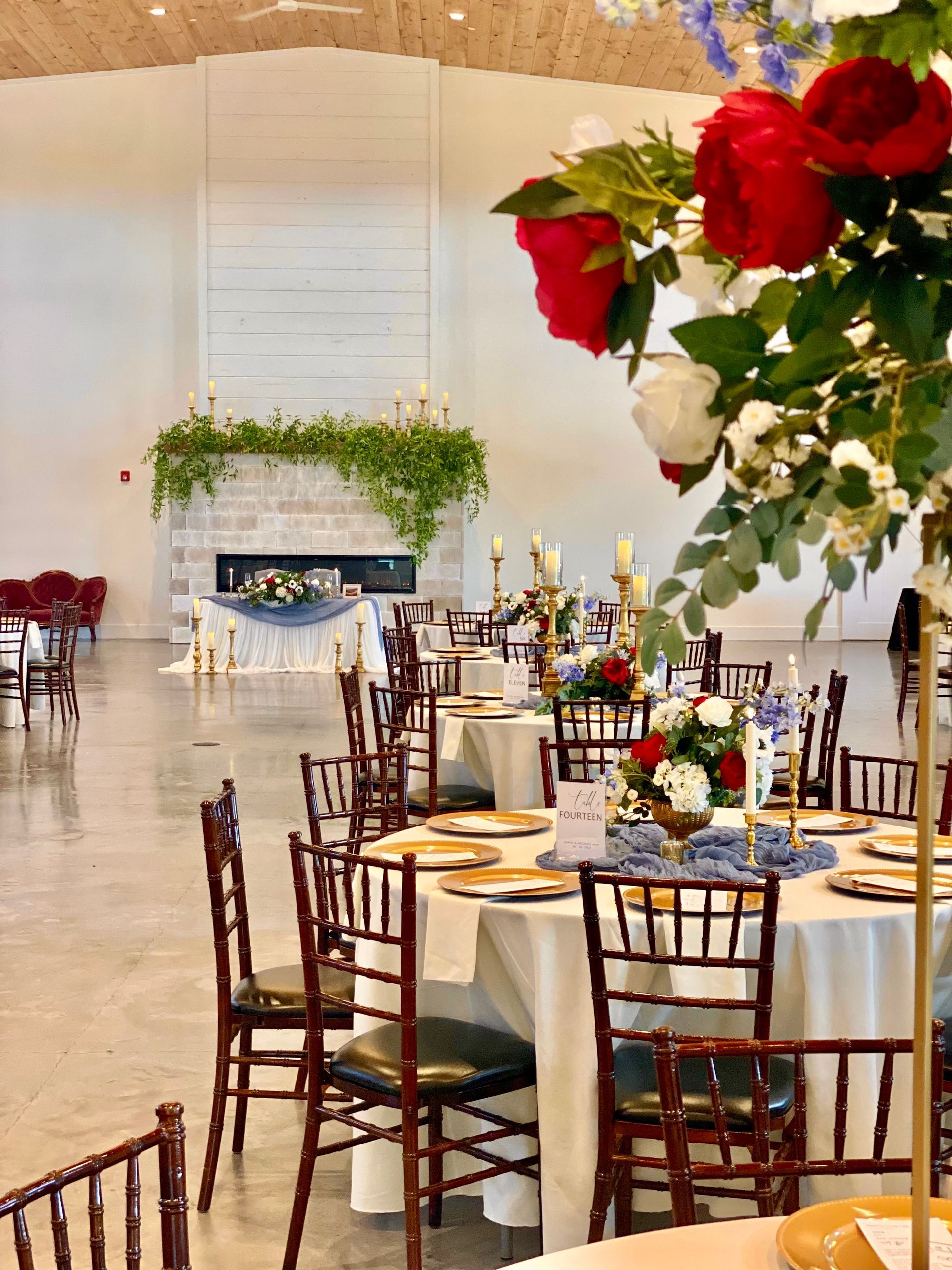 Elegant banquet hall with round tables, gold chairs, and red floral centerpieces beneath a white archway