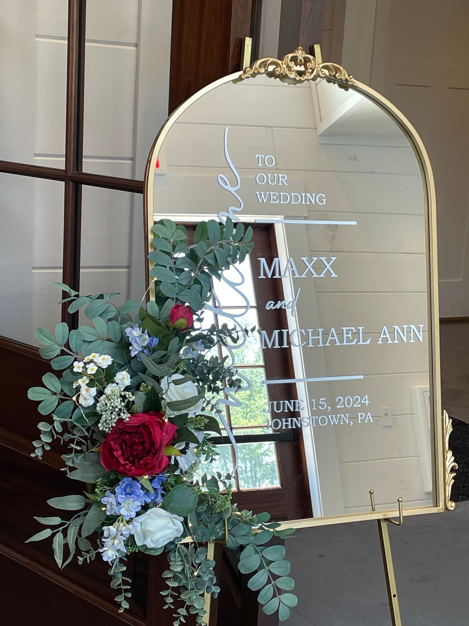 Decorative wedding sign with white text, greenery, and red and white flowers on a gold arch mirror