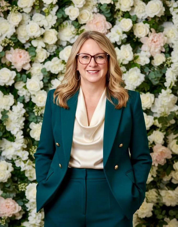 Woman in a teal suit and cream blouse standing before a white floral wall, smiling.