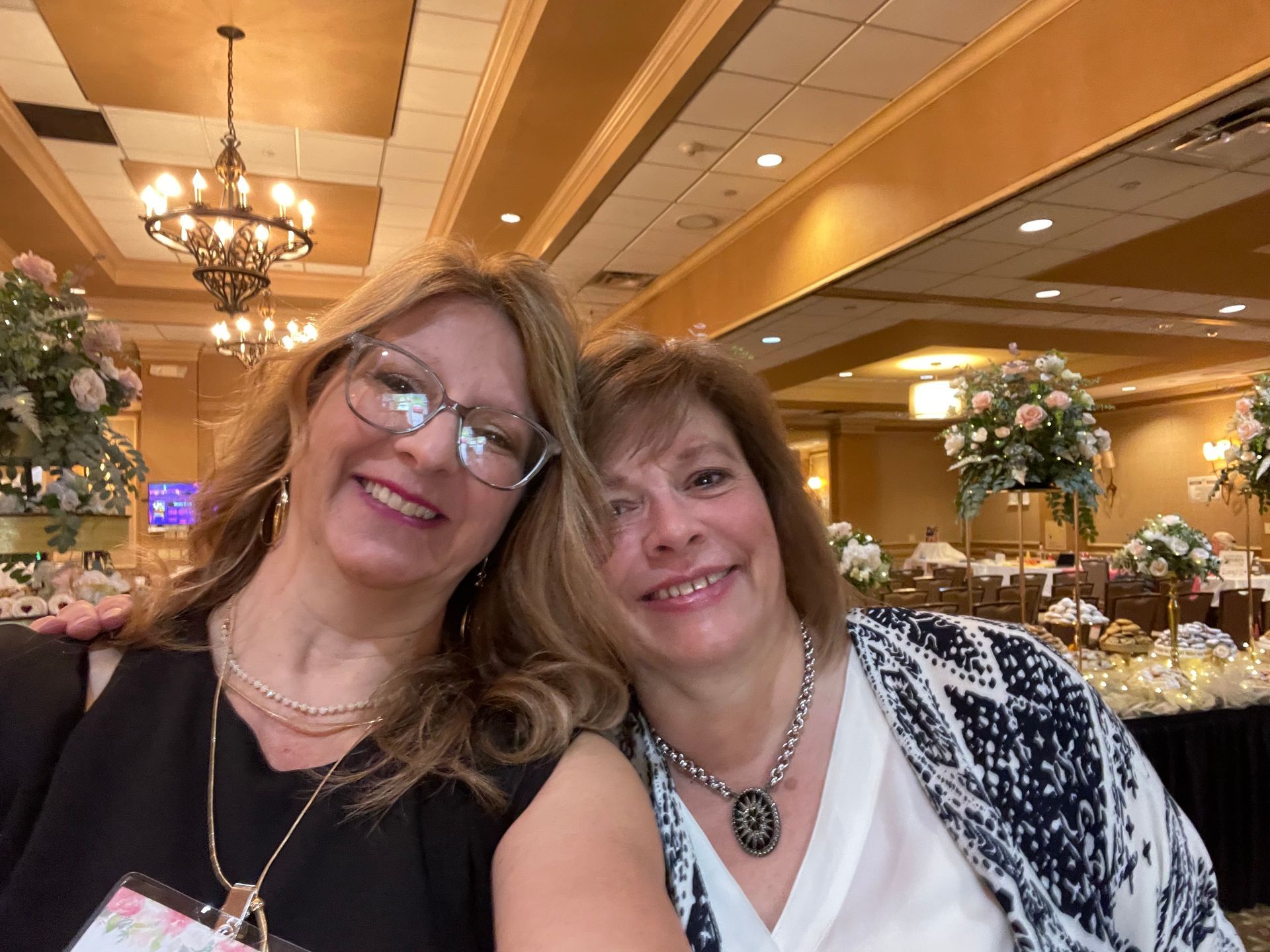 Two smiling people posing together in a warmly lit banquet hall with chandeliers and floral centerpieces