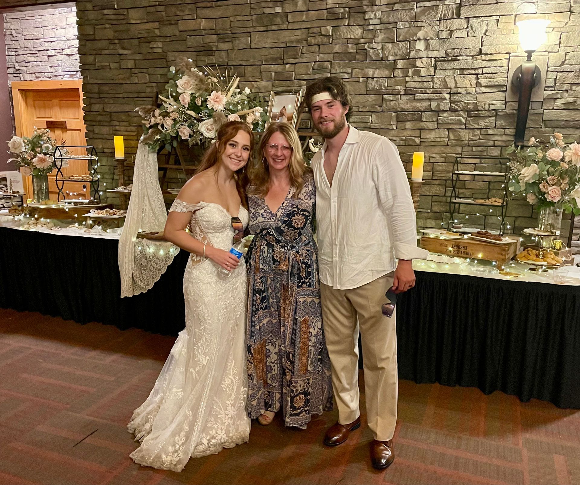 Three people posing at a decorated indoor event with flowers, candles, and a stone wall backdrop.