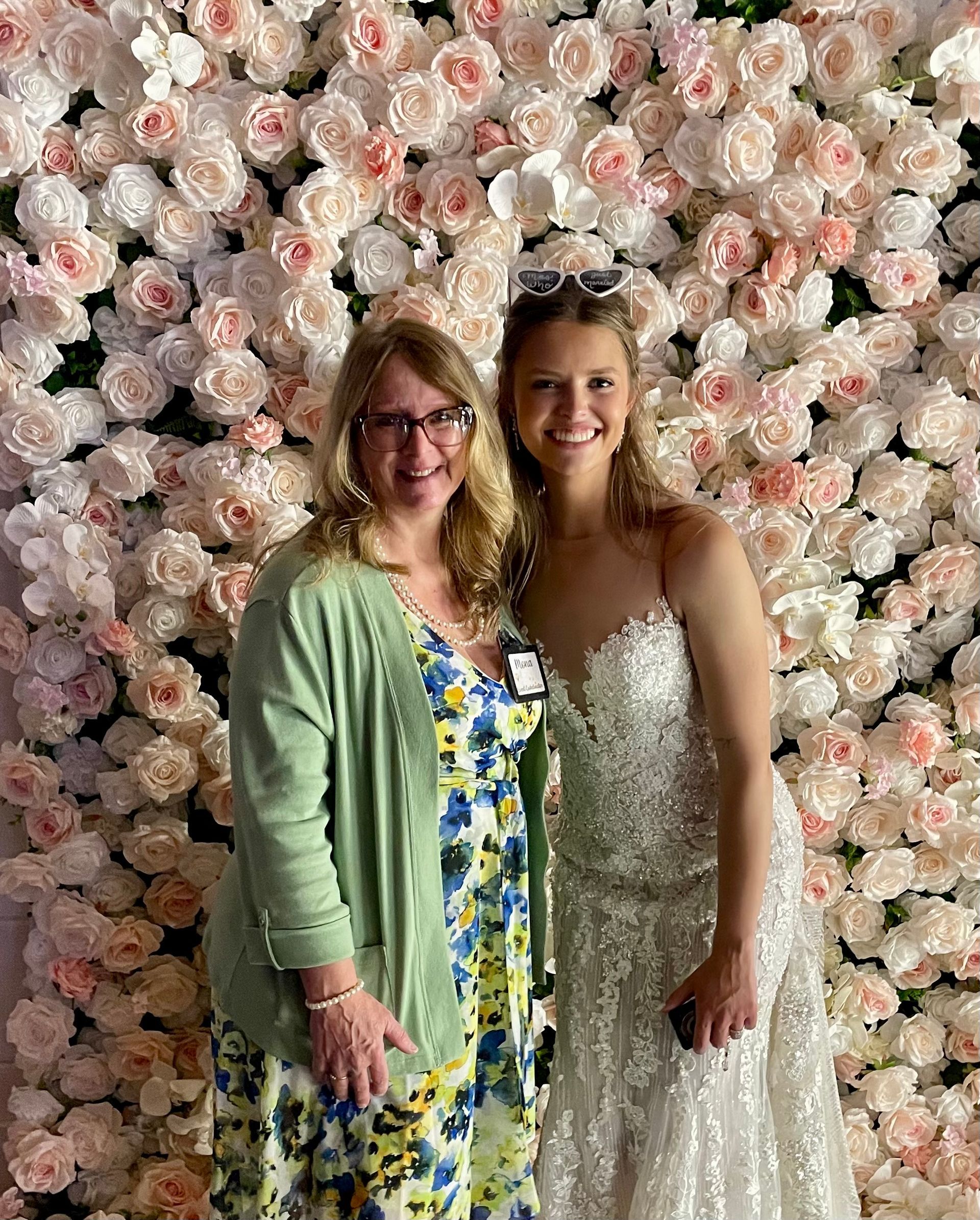 Two women standing together in front of a pink-and-white flower wall, smiling at the camera.