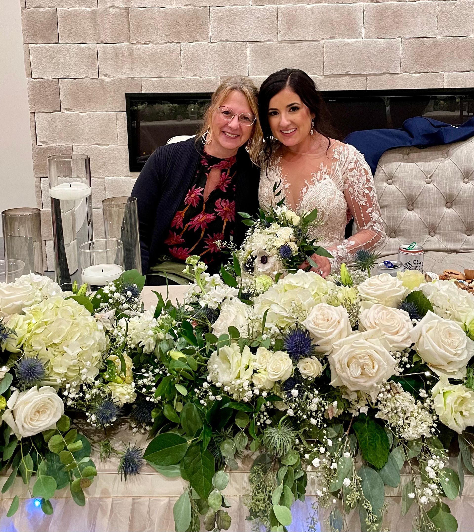 Two women seated behind a white floral arrangement, smiling in front of a stone fireplace.