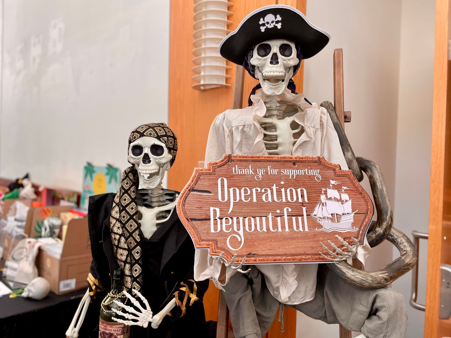 Two skeleton figures holding a wooden “Operation Begointh” sign in a decorated indoor setting