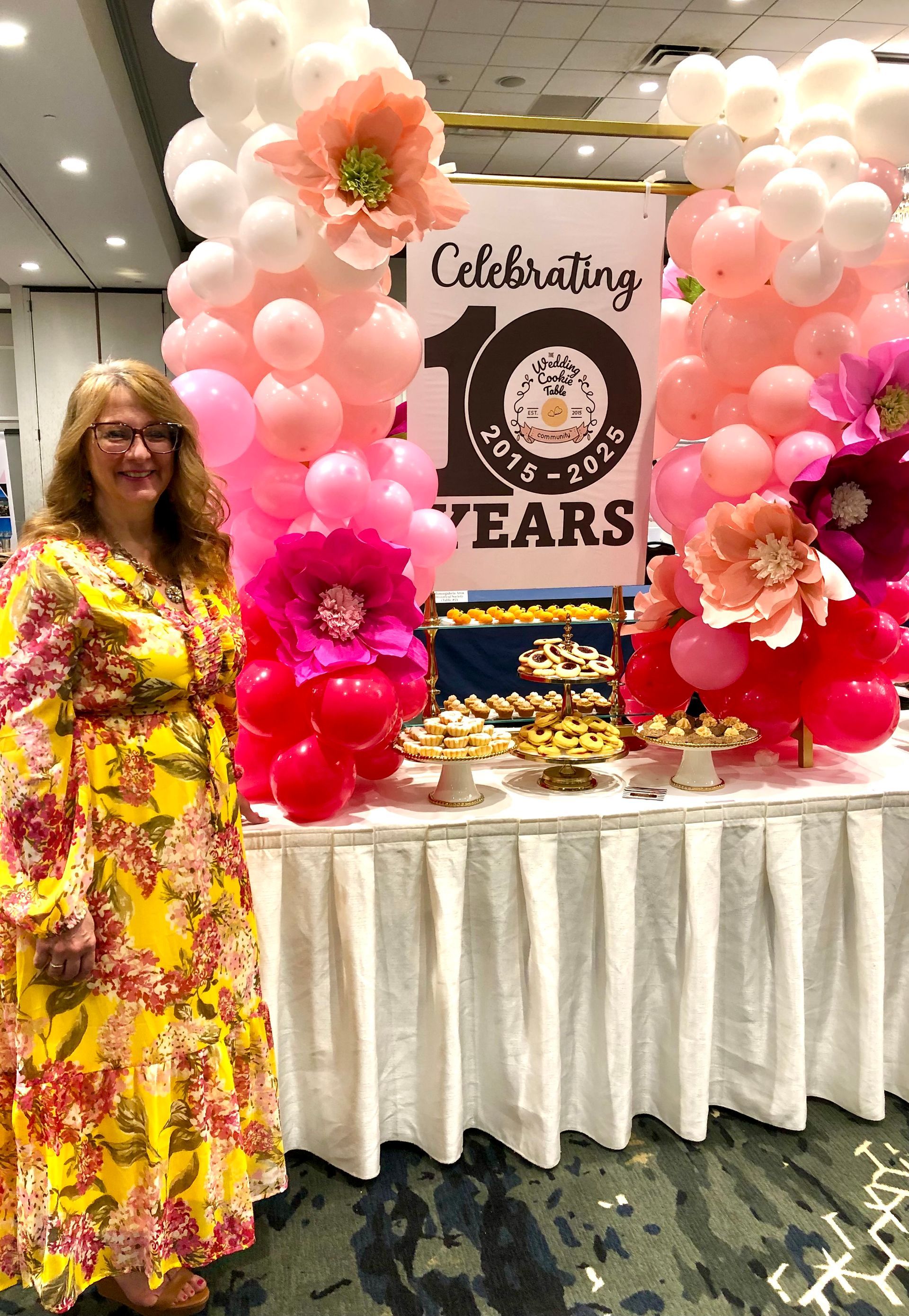 Woman in floral dress standing beside a pink anniversary celebration display and dessert table