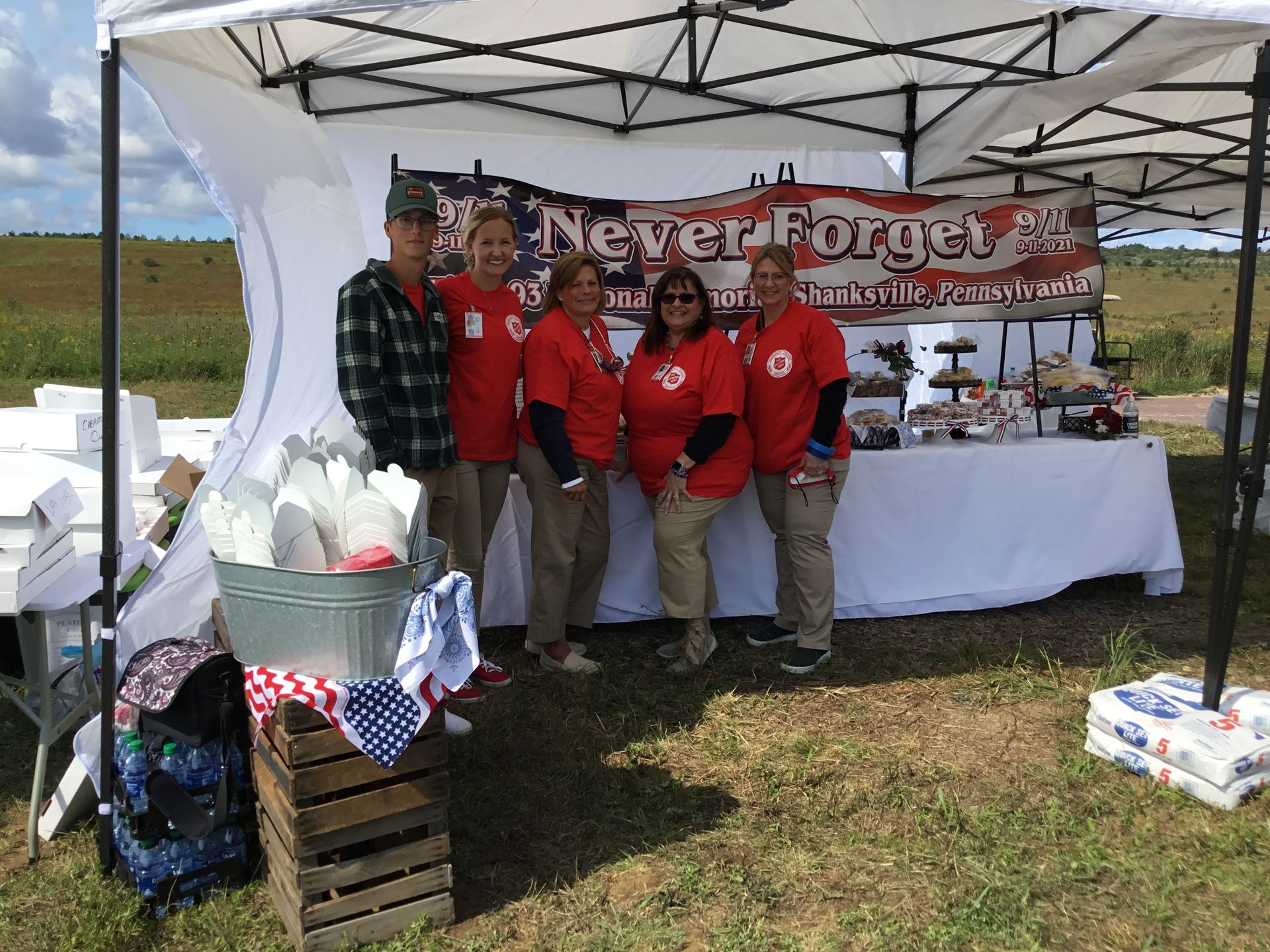 Group of volunteers in red shirts at a booth under a white tent with a “Never Forget” banner.