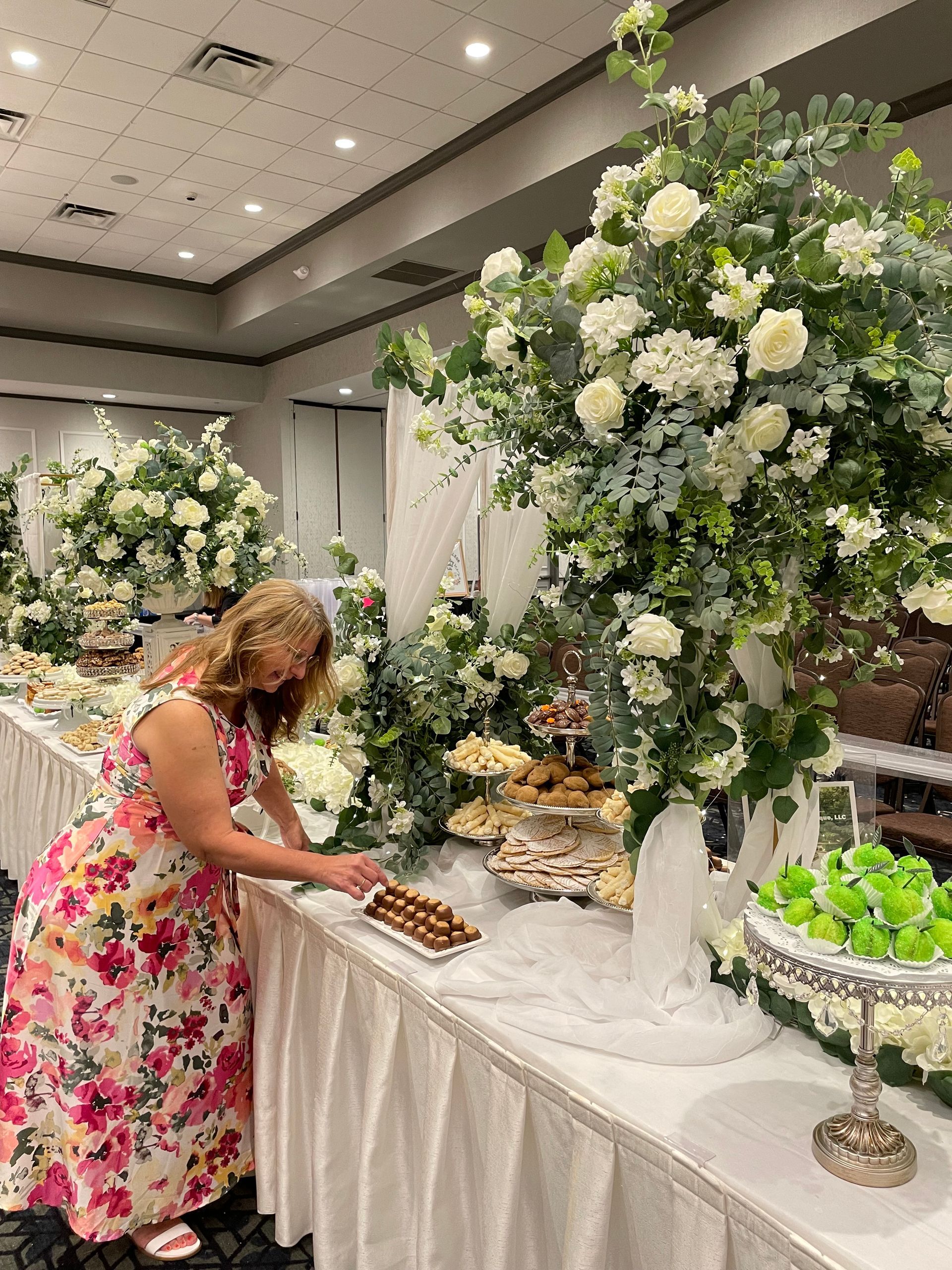Woman in a pink floral dress arranging pastries at a white dessert table with large white flower displays.