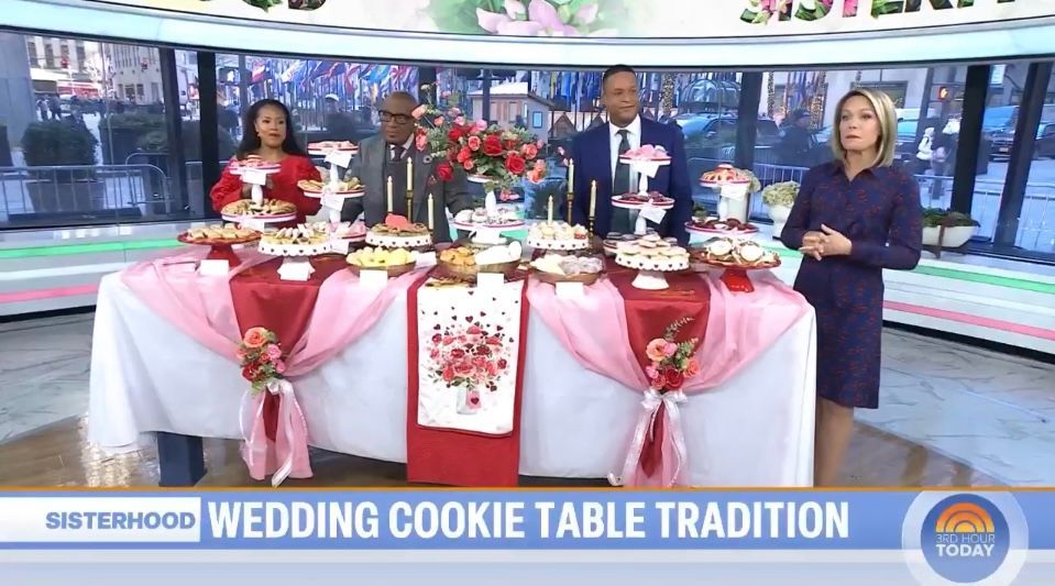 Three people presenting a decorated cookie table on a TV studio set, with pink tablecloths and red accents.