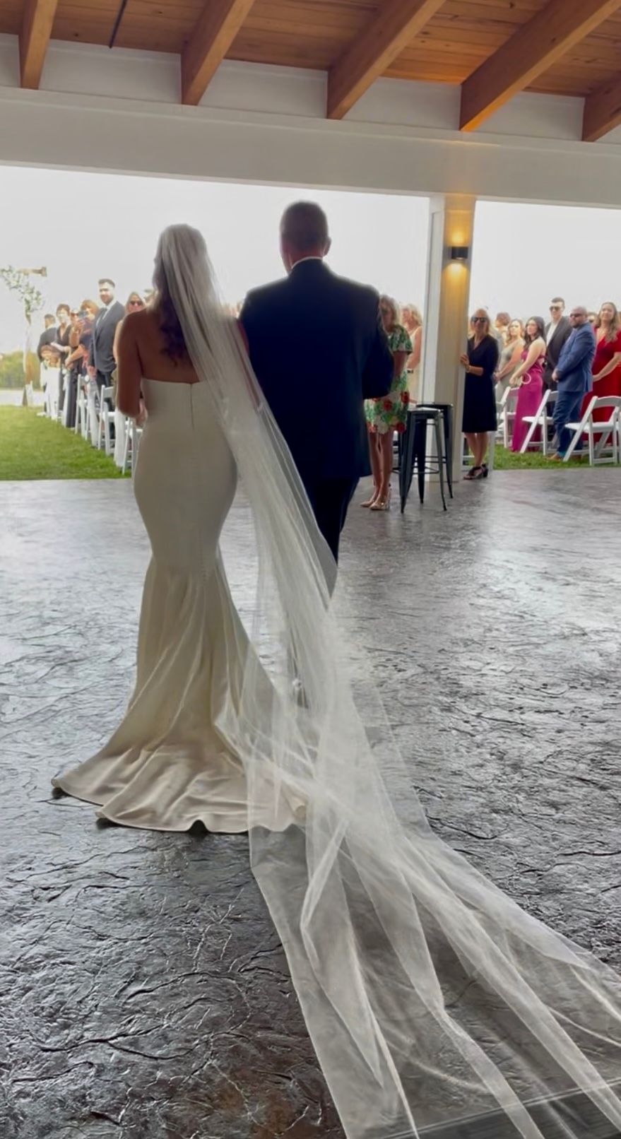 Bride in white gown with long veil walks with groom toward a wedding ceremony indoors
