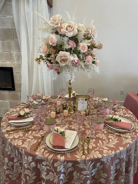 Round banquet table with pink floral decor, gold accents, and a tall rose centerpiece in a bright room