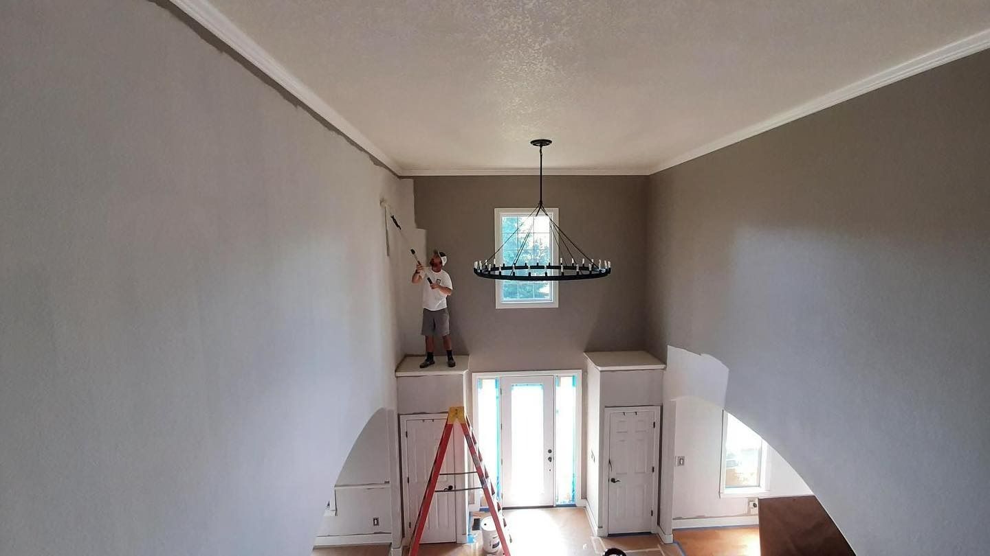 A person on a platform painting a high wall in a room with a chandelier and arched doorway.