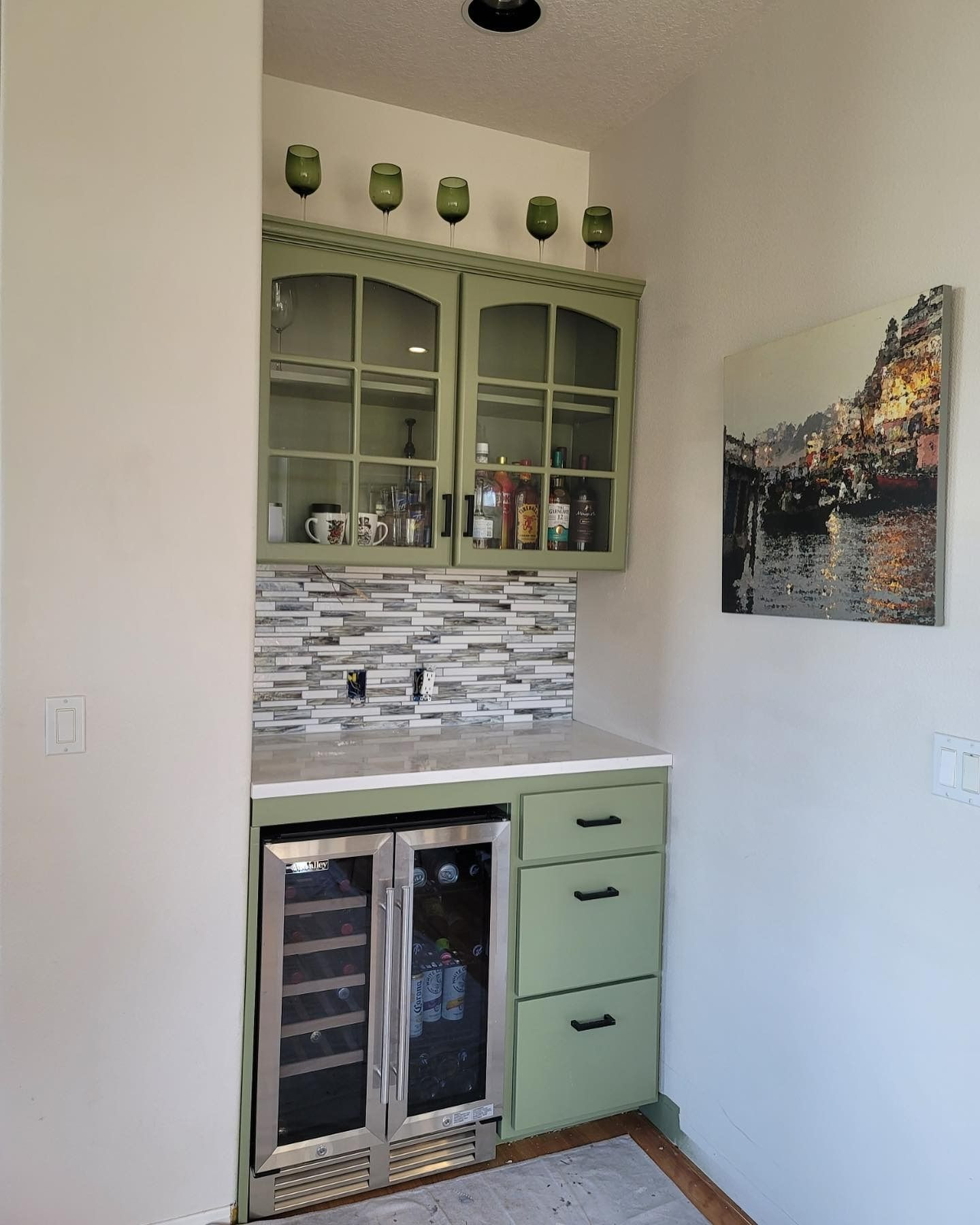 Green-painted home bar with wine fridge, glass-front cabinets, and a tile backsplash.  A canvas painting hangs on the wall.