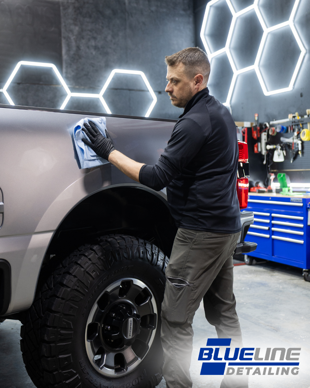 Man wiping a truck's side panel with a microfiber cloth; shop interior with honeycomb lights and tools.