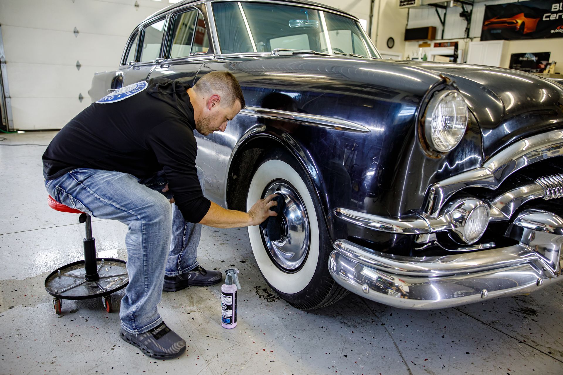 Man cleaning a classic black car's white-walled tire, in a garage.