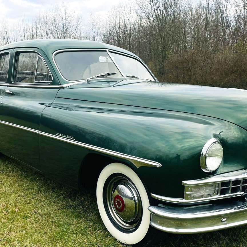 Green vintage Packard sedan with white-wall tires parked on grass, cloudy sky background.