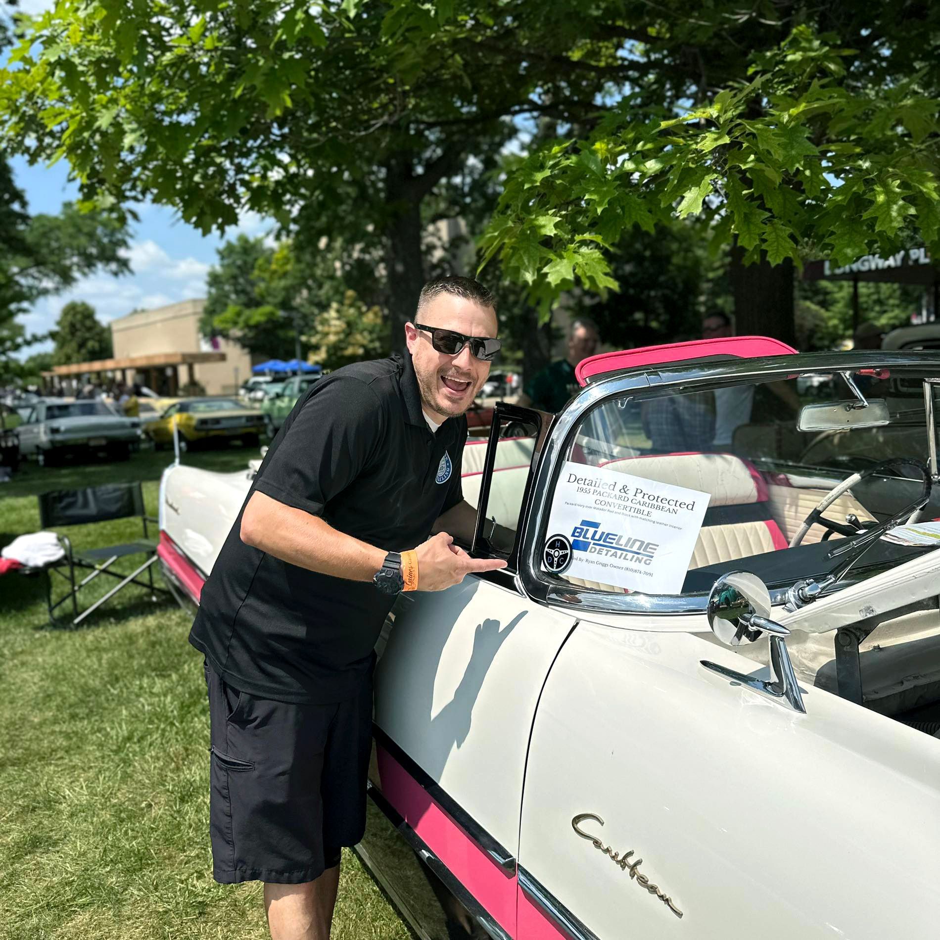 Man points to a white and pink convertible at a car show. He smiles; sunny outdoor setting.