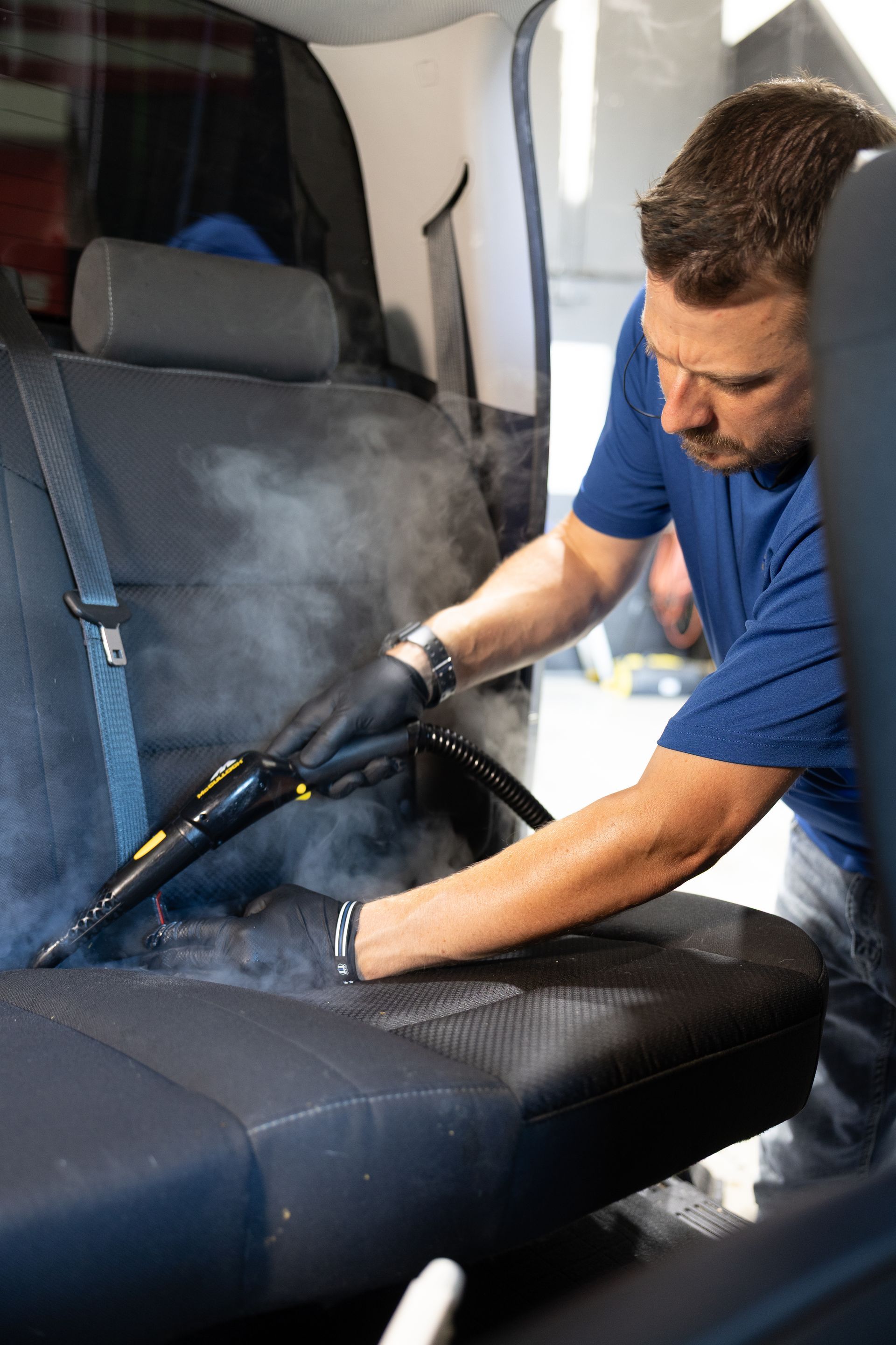 Man steam cleans a black car seat with a steamer, wearing gloves.