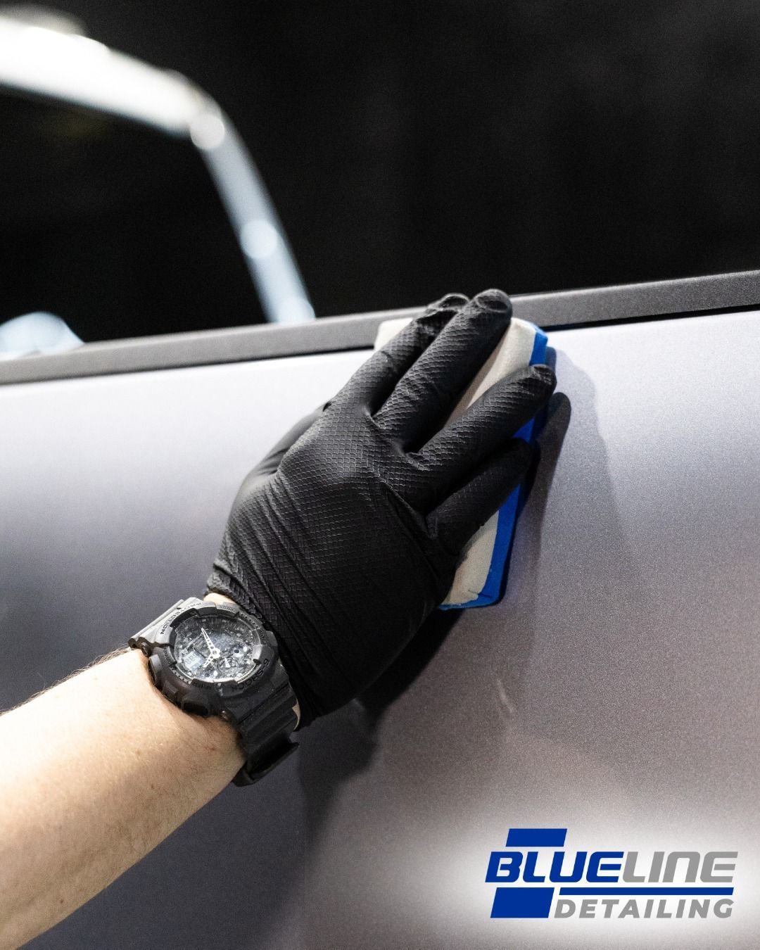 Hand applying detailing product to car with sponge and wearing black glove.