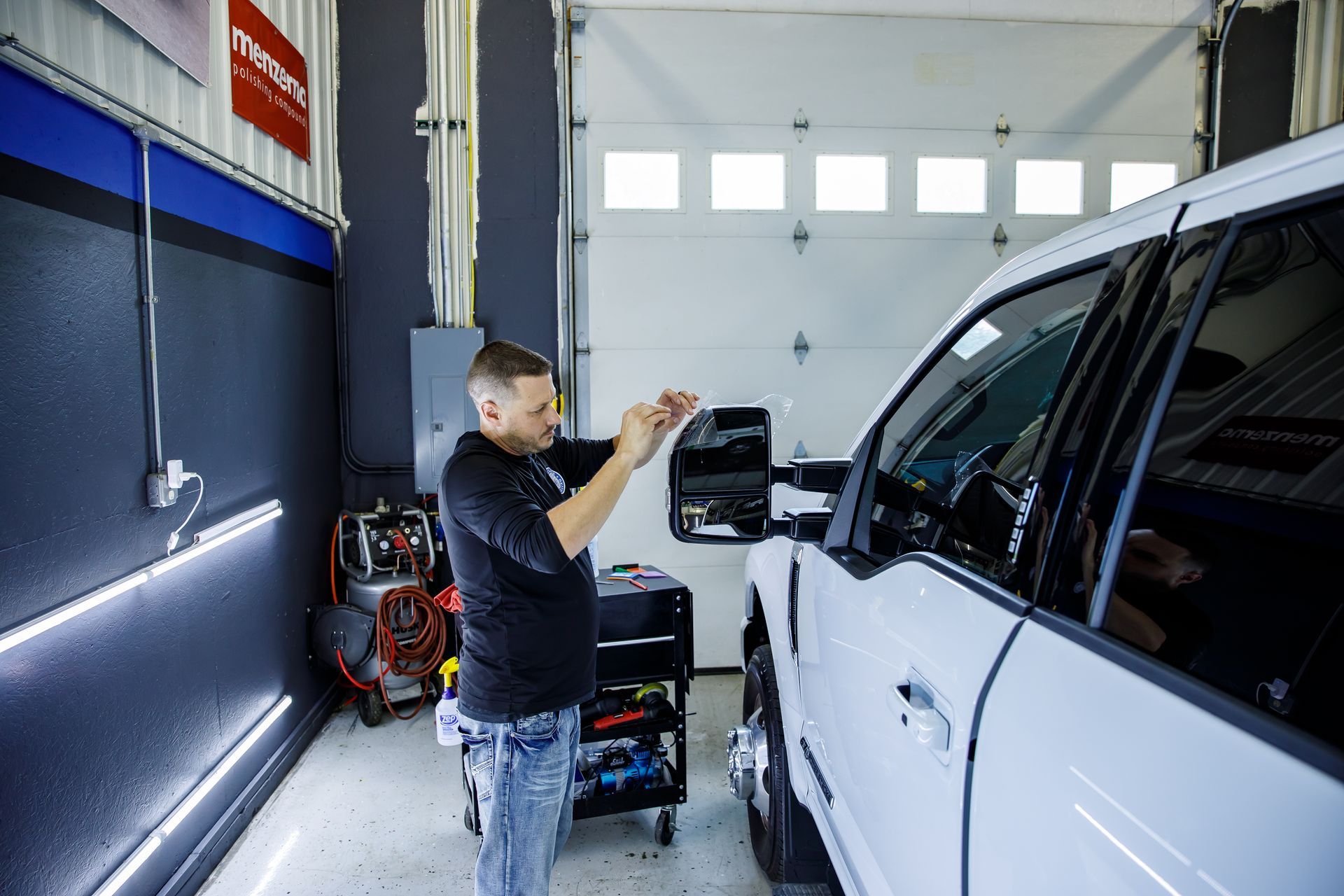 Man working on a white truck's side mirror in a garage.