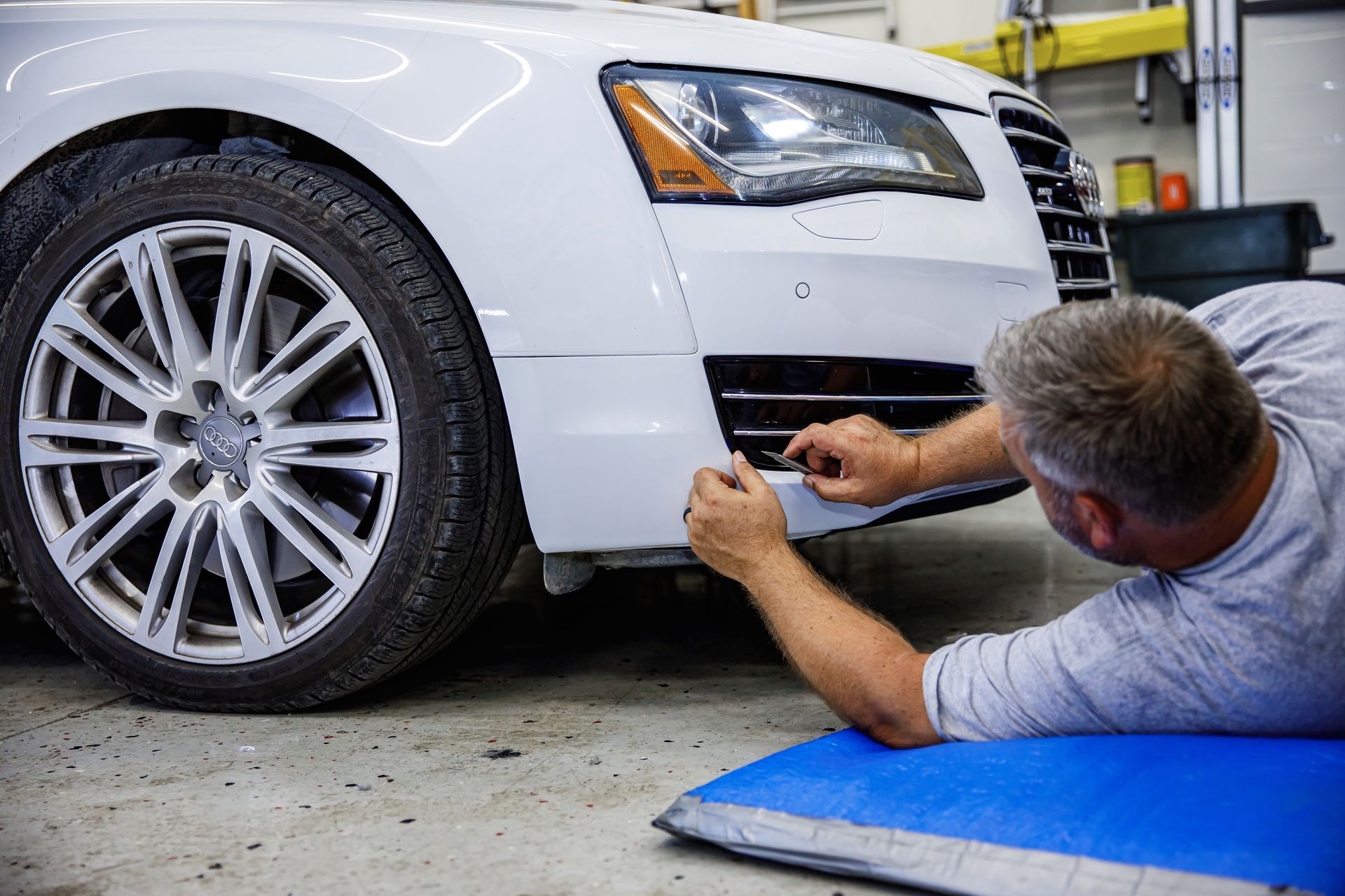 Man repairs white car bumper. He's on a blue mat in a garage.