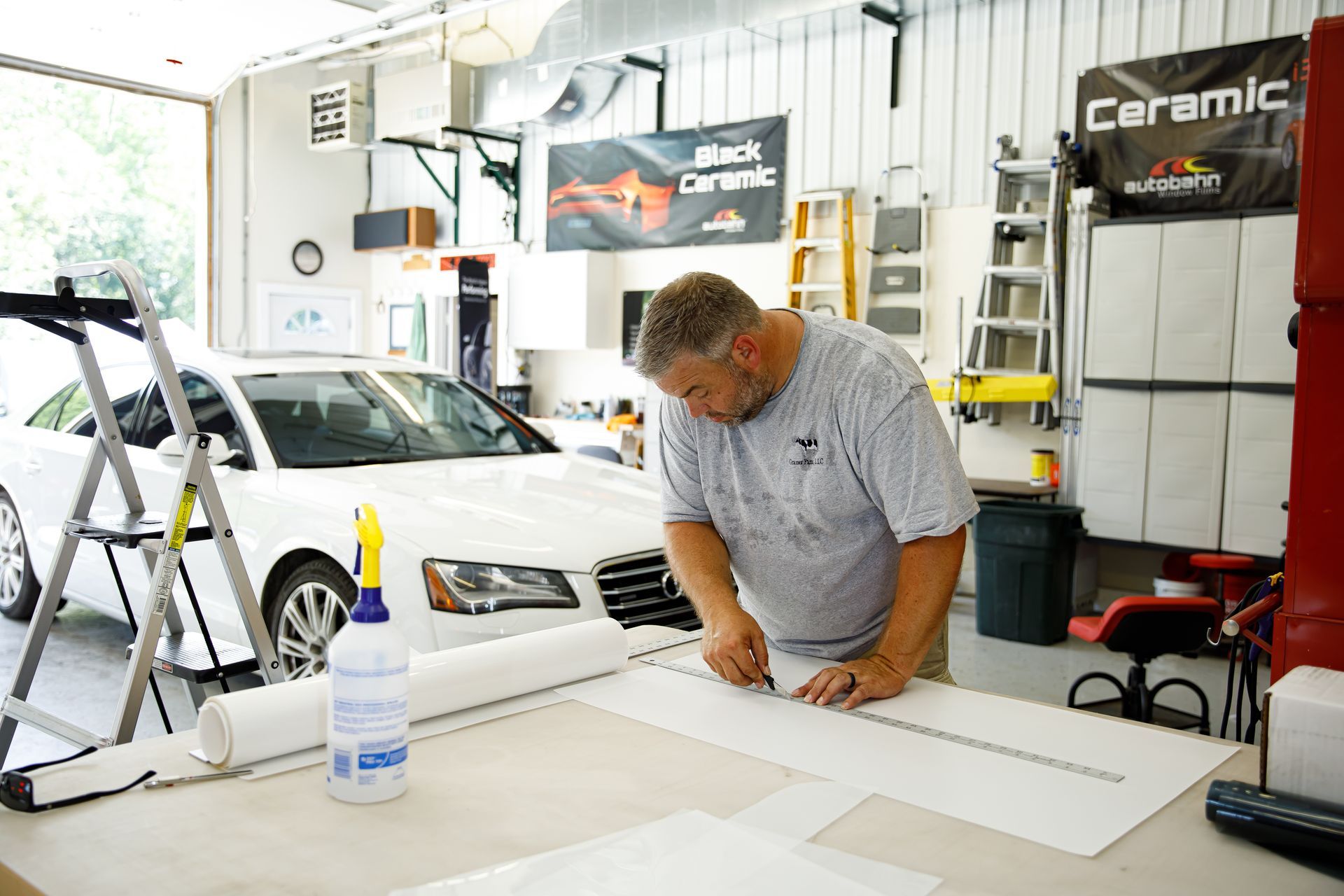 Man cuts material on a table in a garage with a car. Rolls of material, tools and a water bottle are visible.