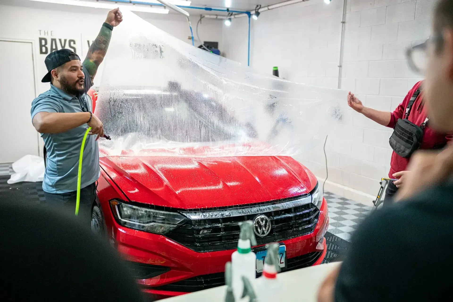 People applying clear film to a red car in a shop. One man sprays water, another holds the film.