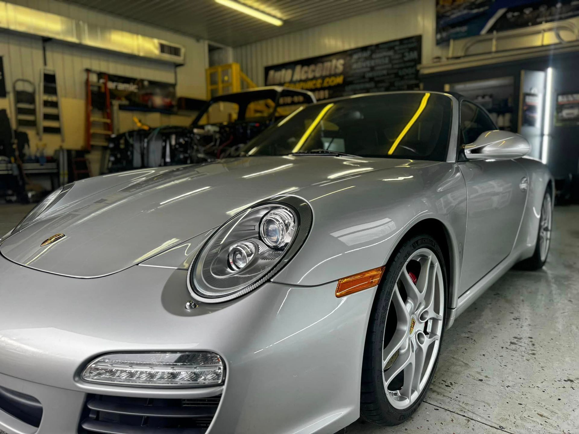 Silver Porsche convertible in a garage, shiny exterior, overhead lighting.
