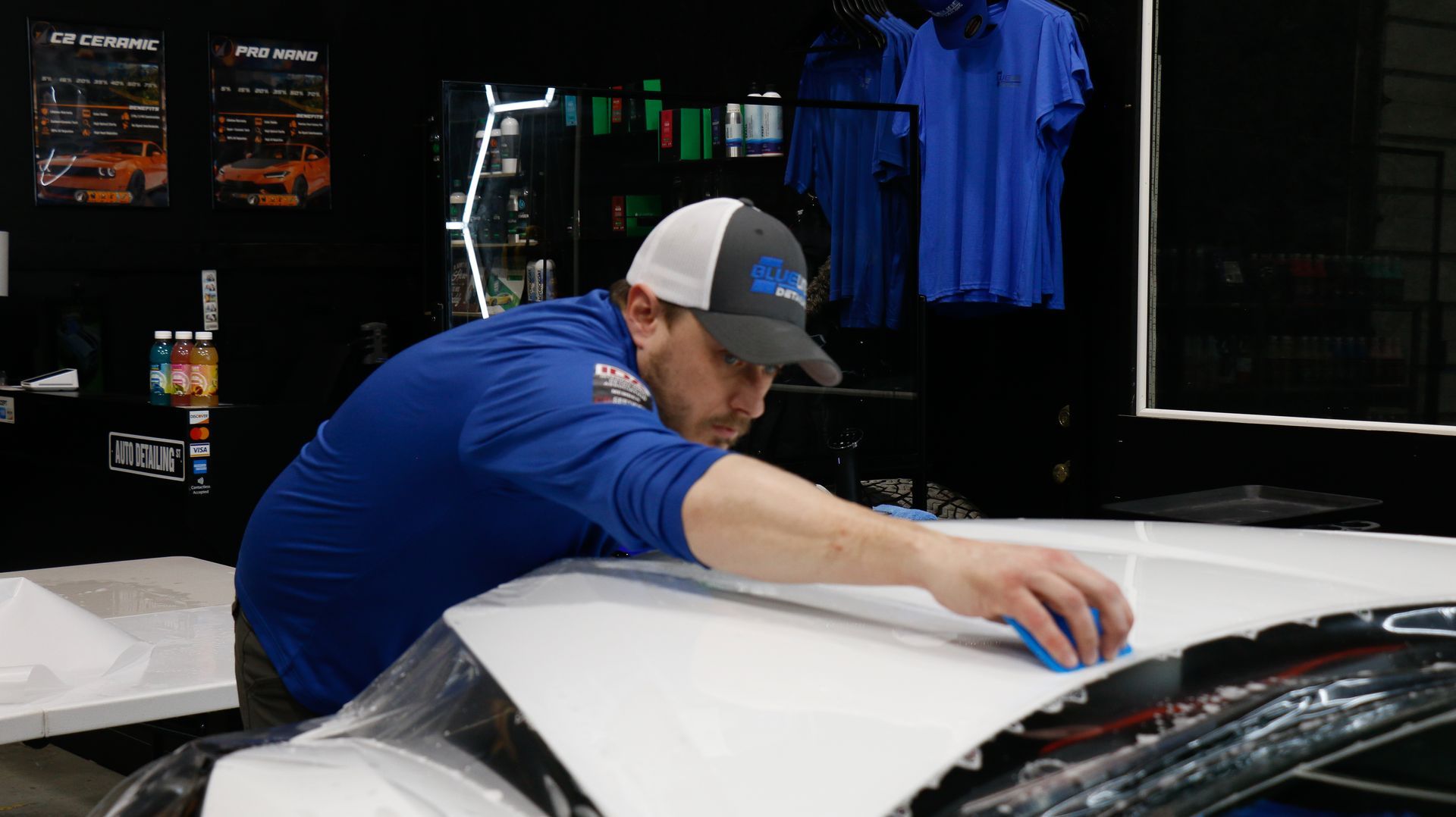 Man in blue shirt and hat applies film to a white car hood. Interior shop with tools and displays in the background.