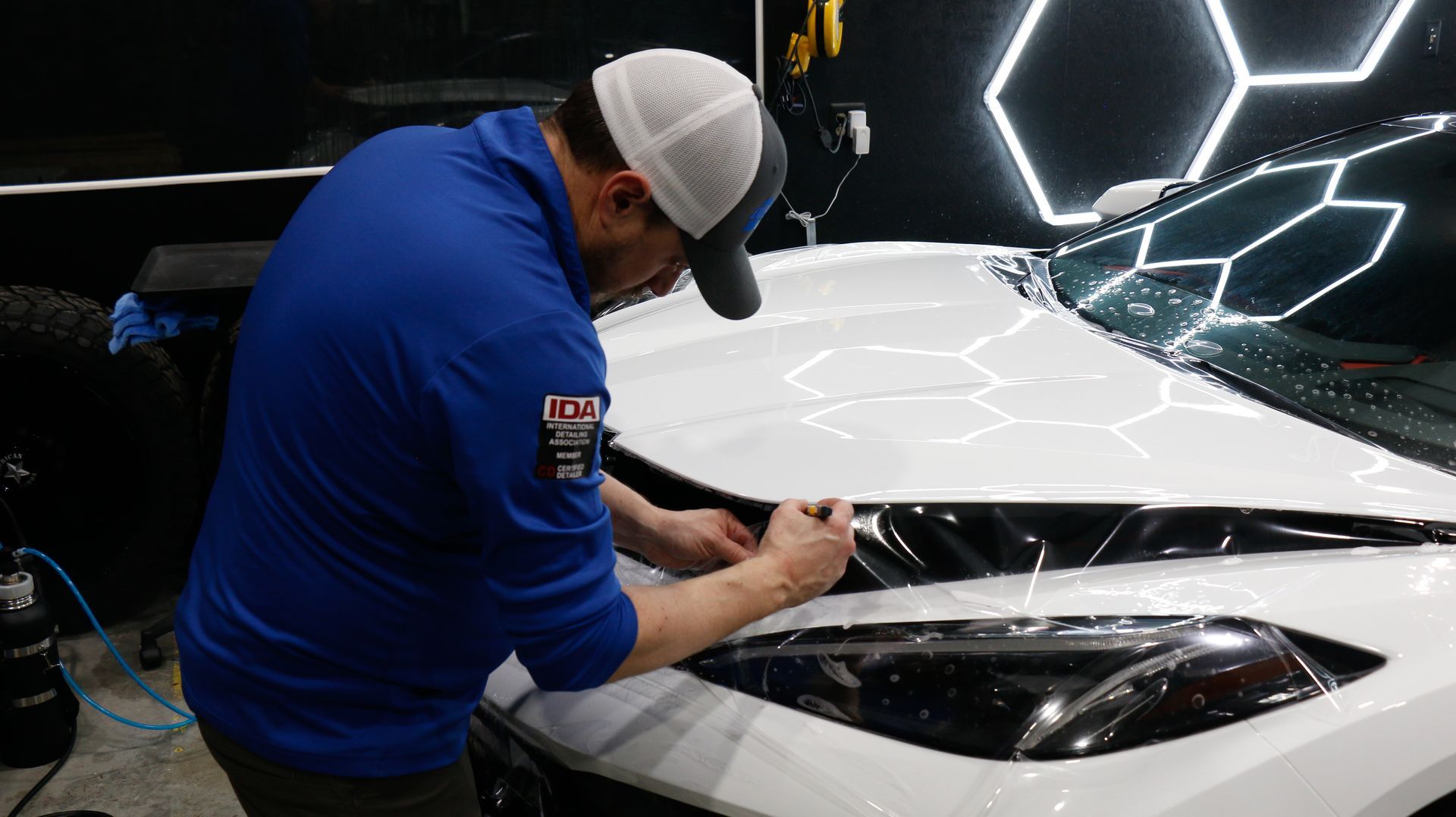 Person applying film to the hood of a white car in a well-lit shop.