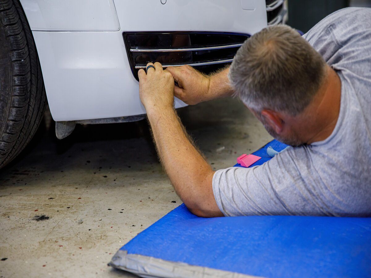 Man installing a part on a white car, kneeling on a blue mat in a garage.