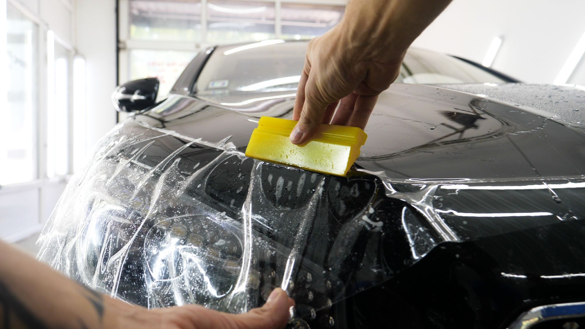Applying clear protective film to a black car's hood with a yellow squeegee inside a garage.