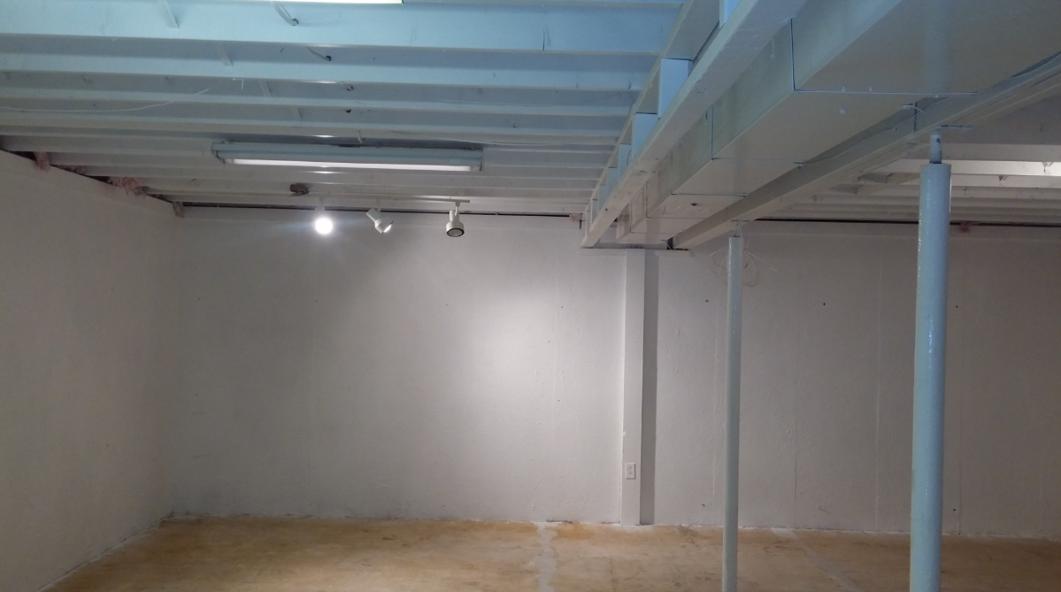 Empty unfinished room with white walls, exposed blue ceiling beams, and concrete floor.